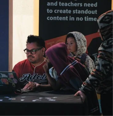 A man with glasses and three young girls gathered around a table, looking at a laptop computer, in an indoor setting with a black banner in the background.