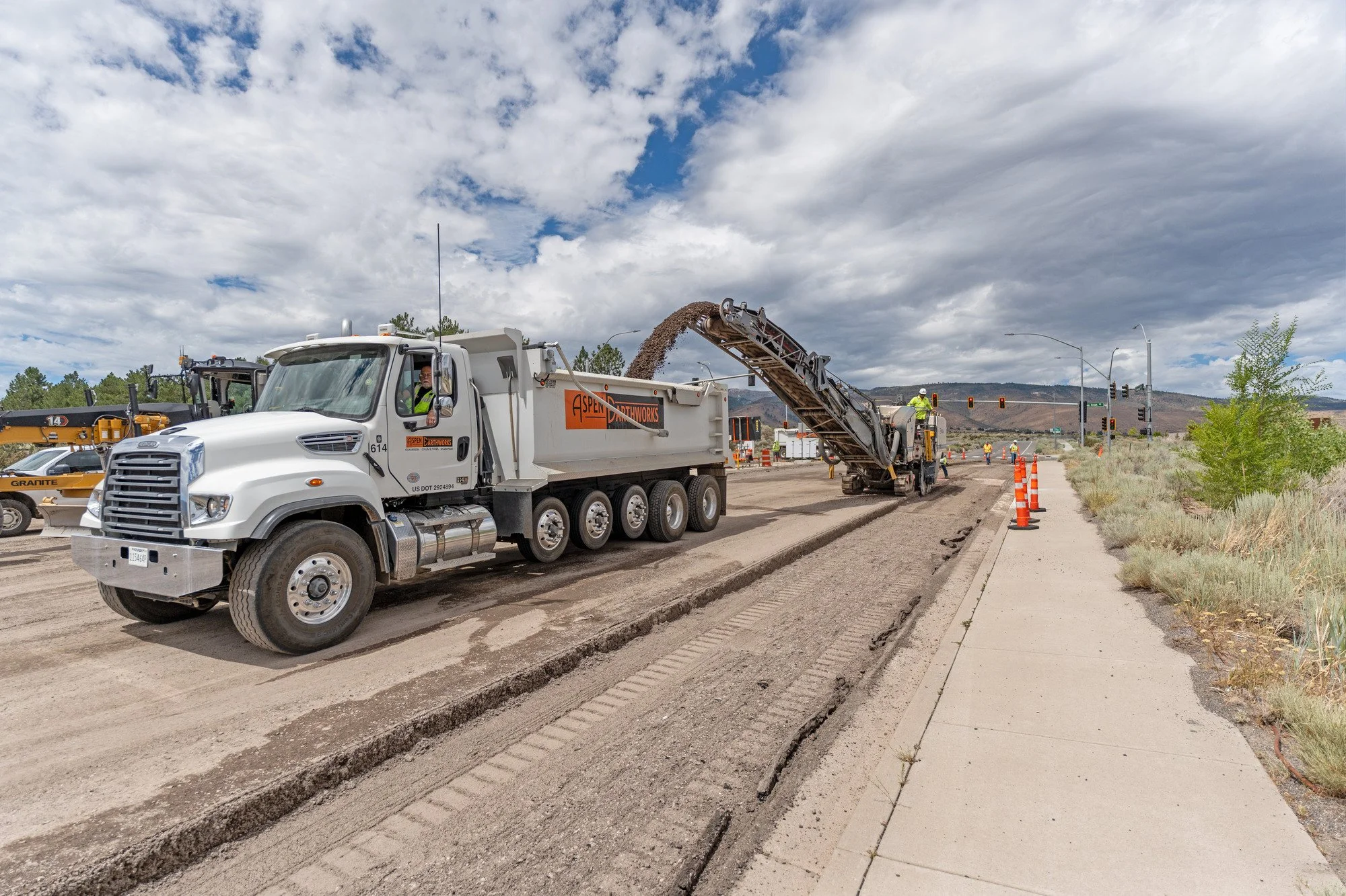 Construction workers use a road milling machine and dump truck on a road under a cloudy sky.