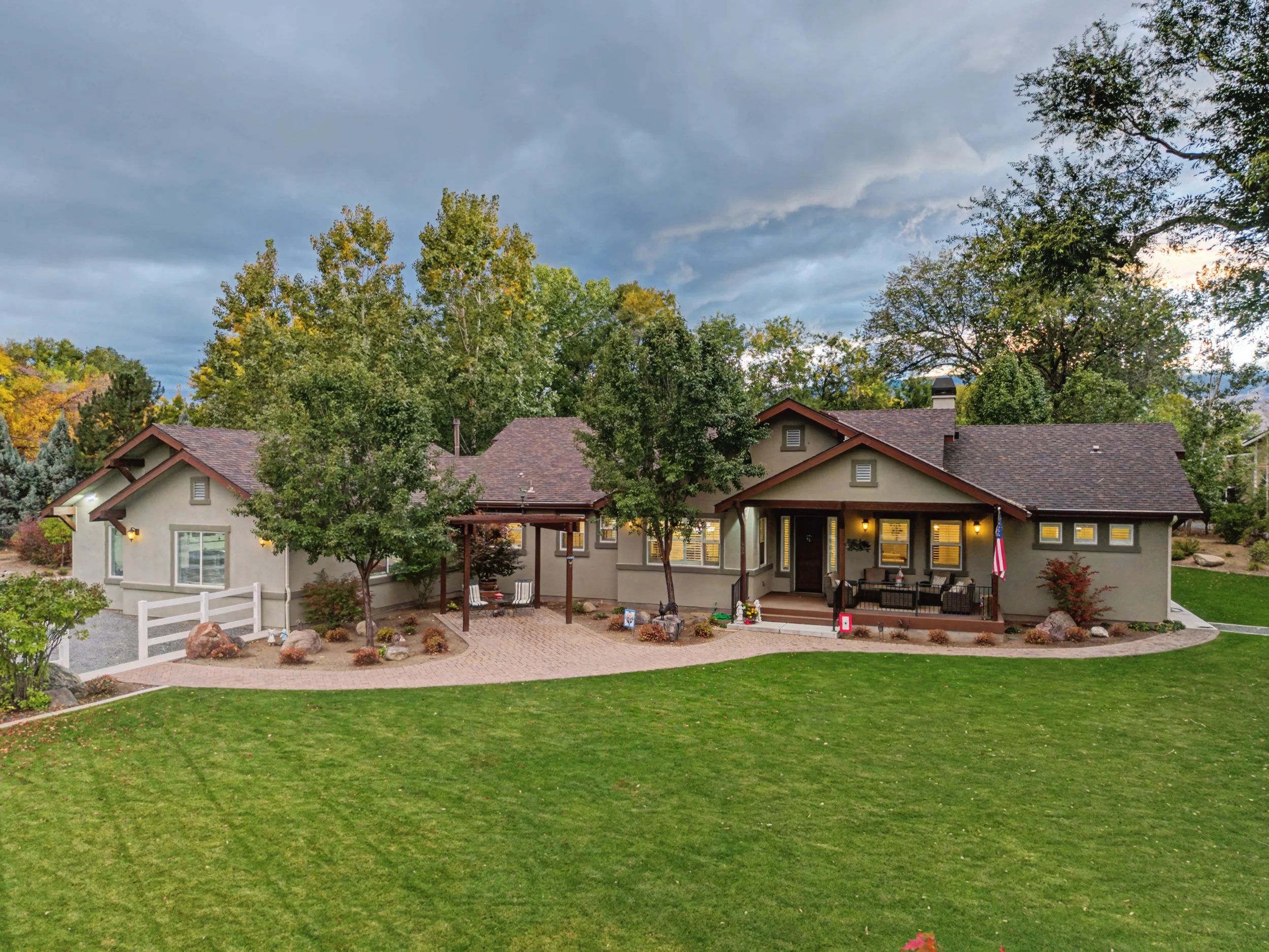 A large suburban house with a well-maintained lawn, trees, and a brick driveway, under a mostly cloudy sky.