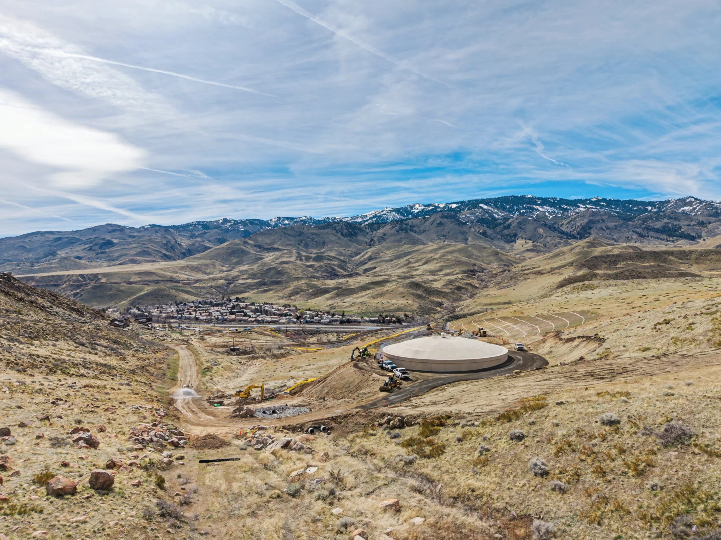 Construction site in a mountainous landscape with a large circular structure, construction vehicles, and a distant small town, under a blue sky with wispy clouds.