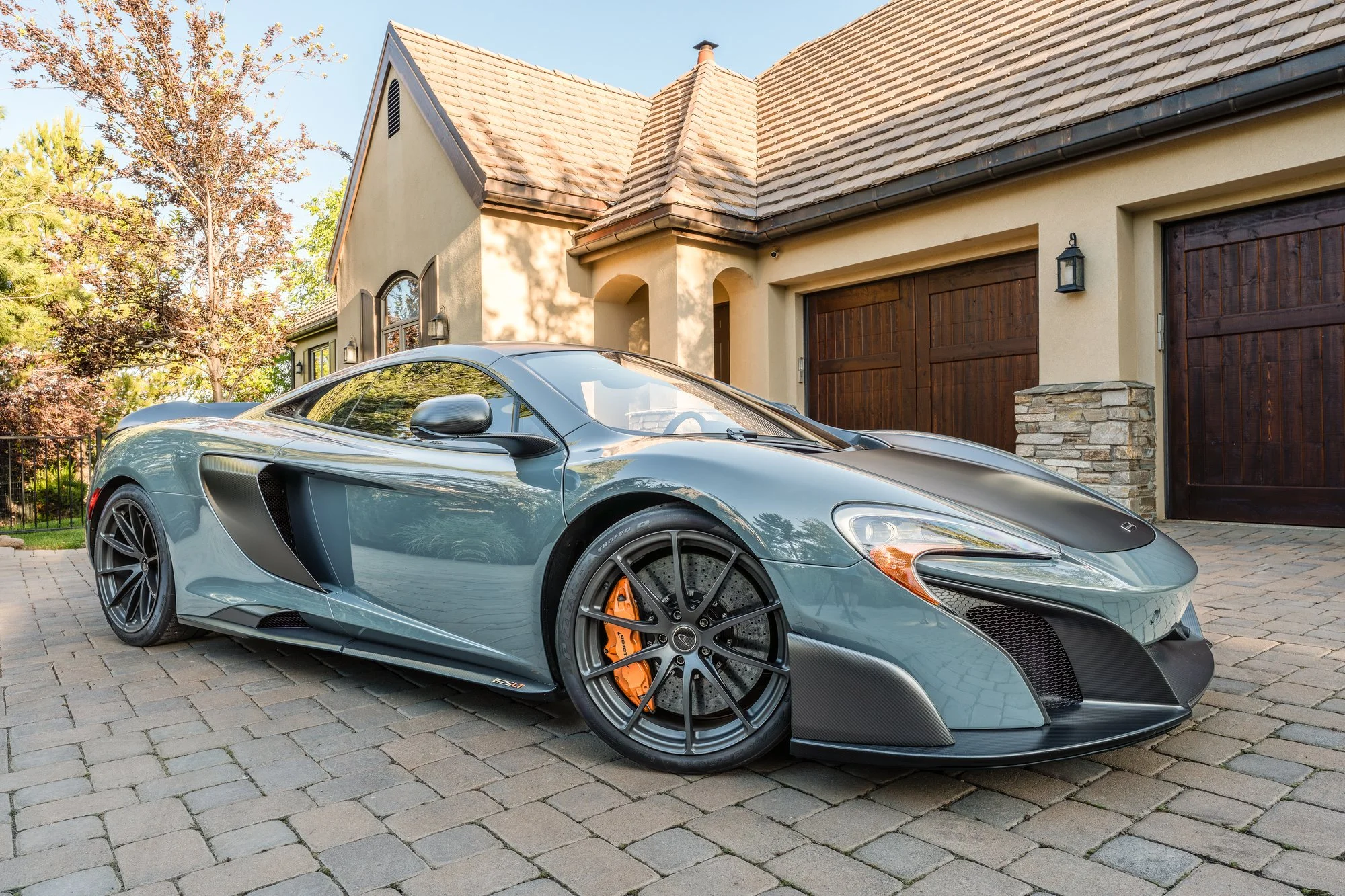A sleek gray sports car parked on a cobblestone driveway in front of a beige house with wooden garage doors and stone accents, surrounded by trees.