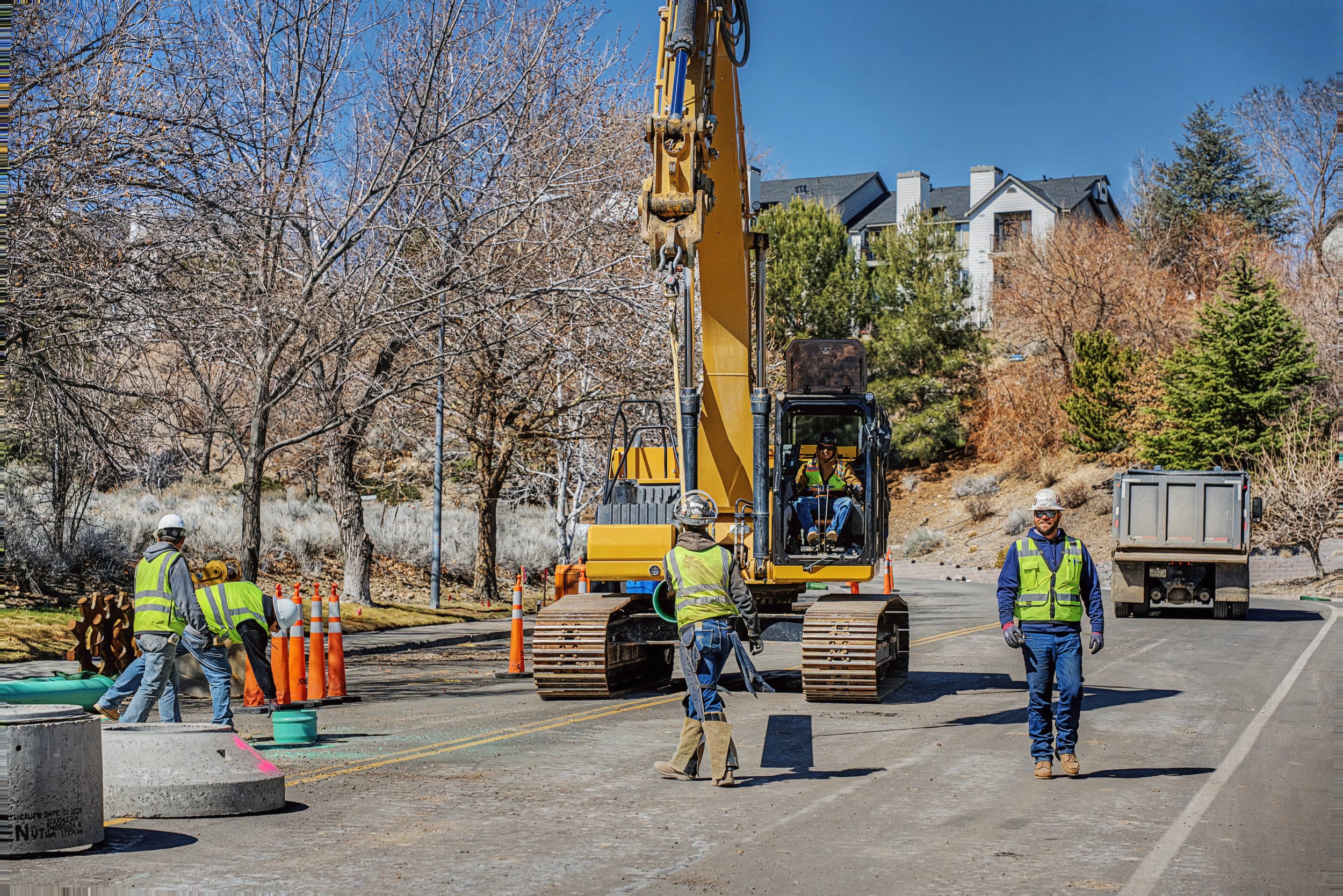 Construction workers operating equipment and working on a street project, with a large excavator and traffic cones, in a residential area with trees and houses in the background.