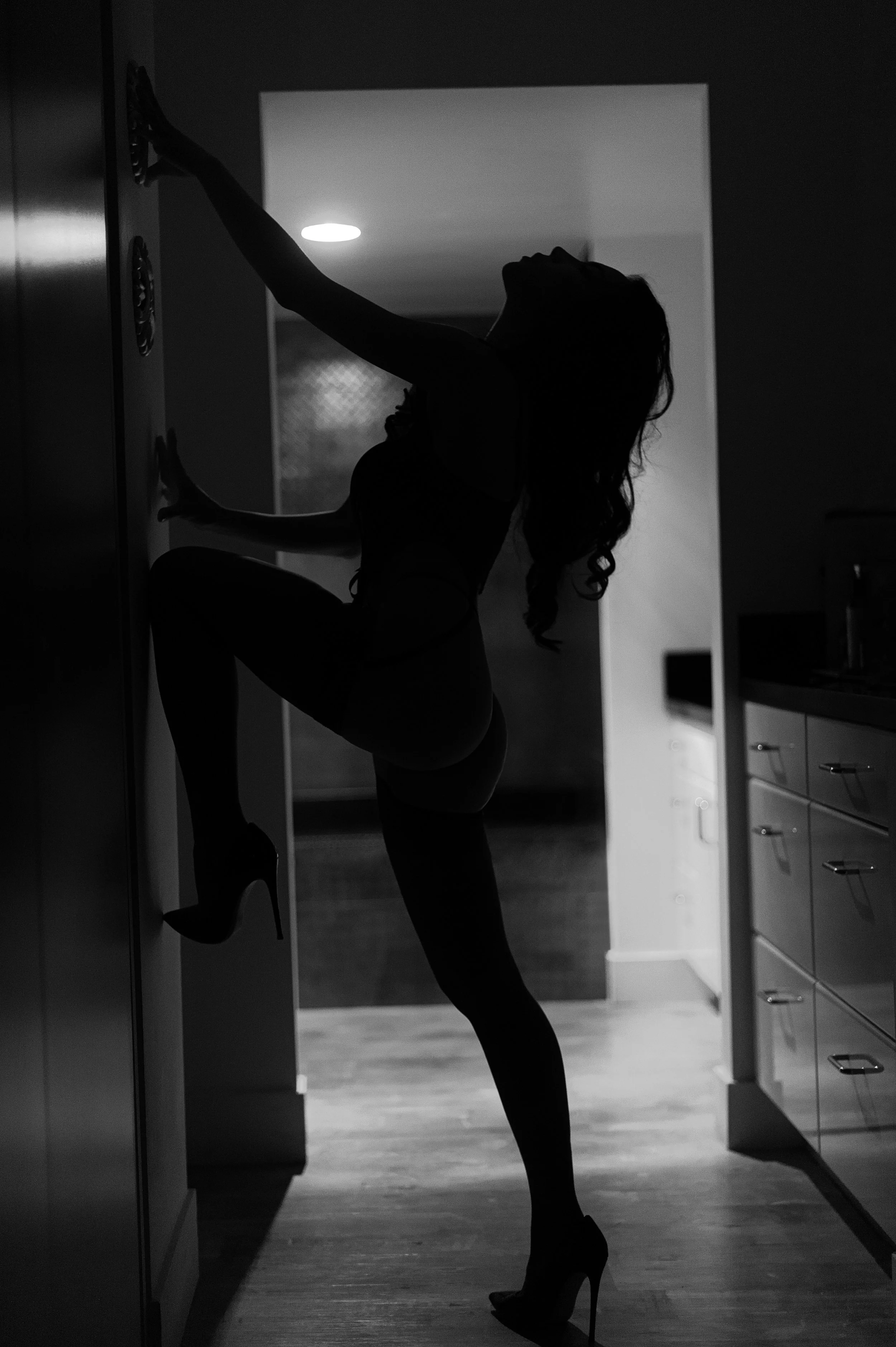 Silhouette of a woman in high heels leaning against a wall in a kitchen, with one leg raised and bent, creating a dramatic and artistic pose. GFE Las Vegas, dinner date, escort, Scarlett Song Escort,