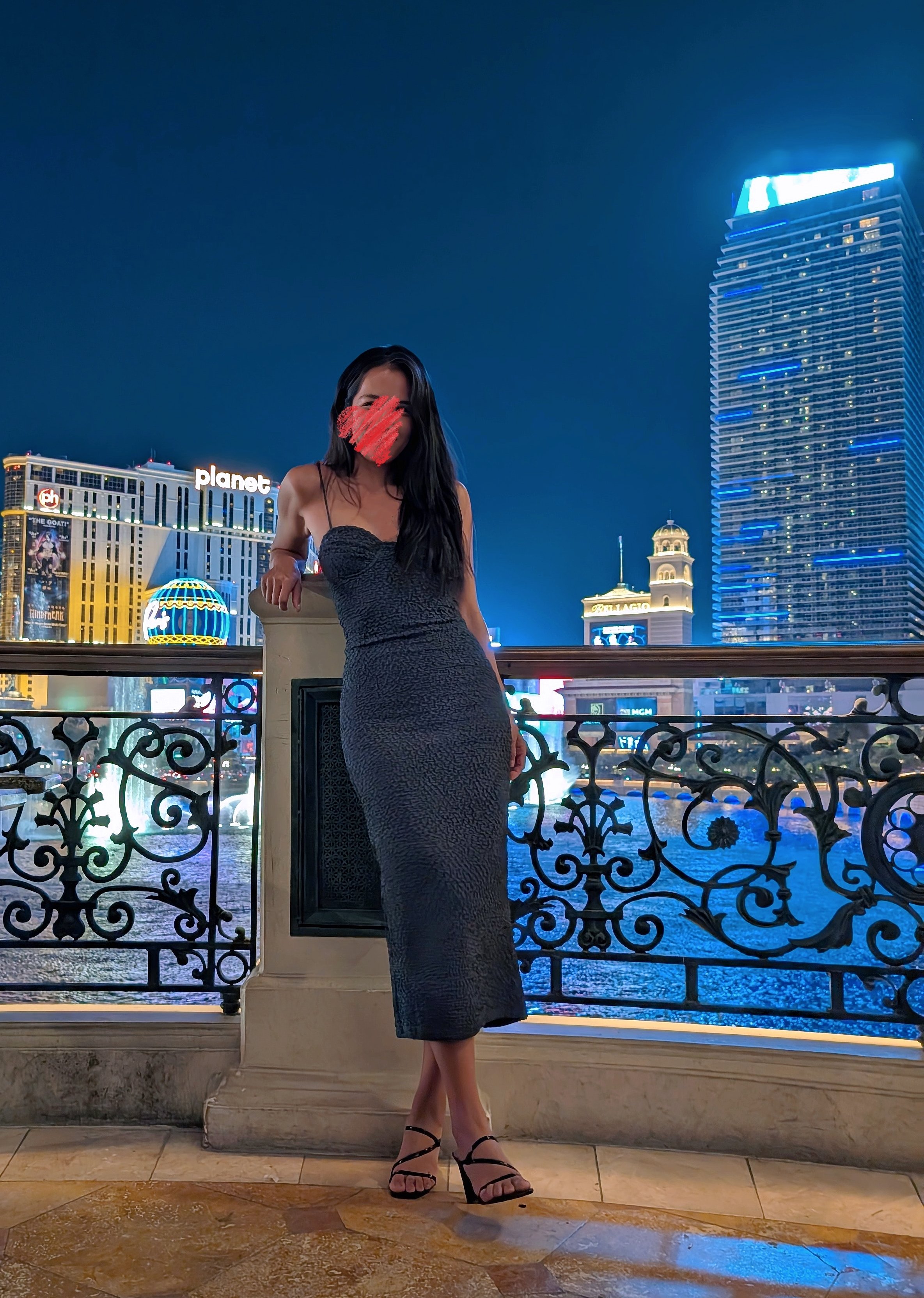 Woman in a black dress and heels posing on a balcony at night with a city skyline, illuminated buildings, and water in the background. GFE Las Vegas, dinner date, escort, Scarlett Song Escort,