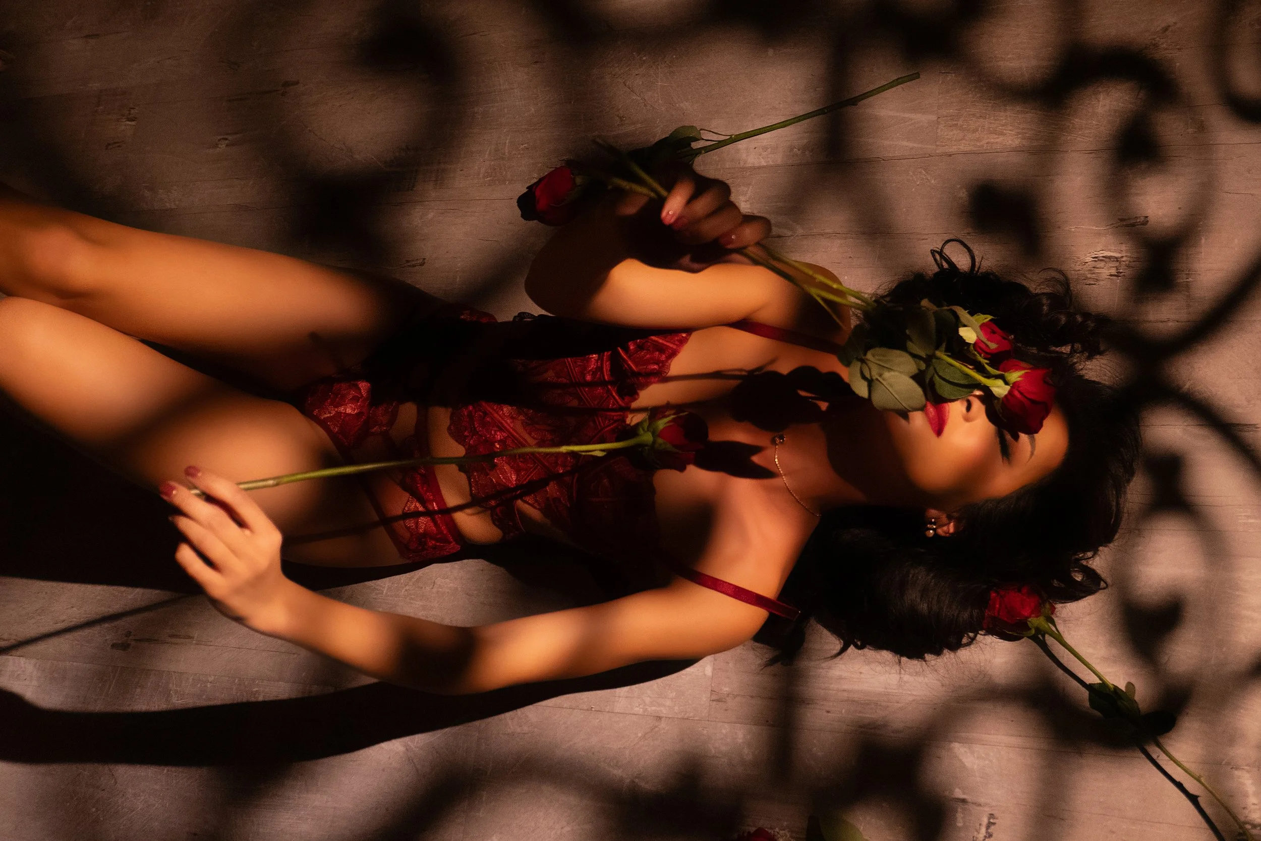 A woman with dark, curly hair is lying on a wooden floor, holding red roses in her hands. She is wearing red lace lingerie and a necklace, partially covered by shadows and leaves. GFE Las Vegas, dinner date, escort, Scarlett Song Escort,