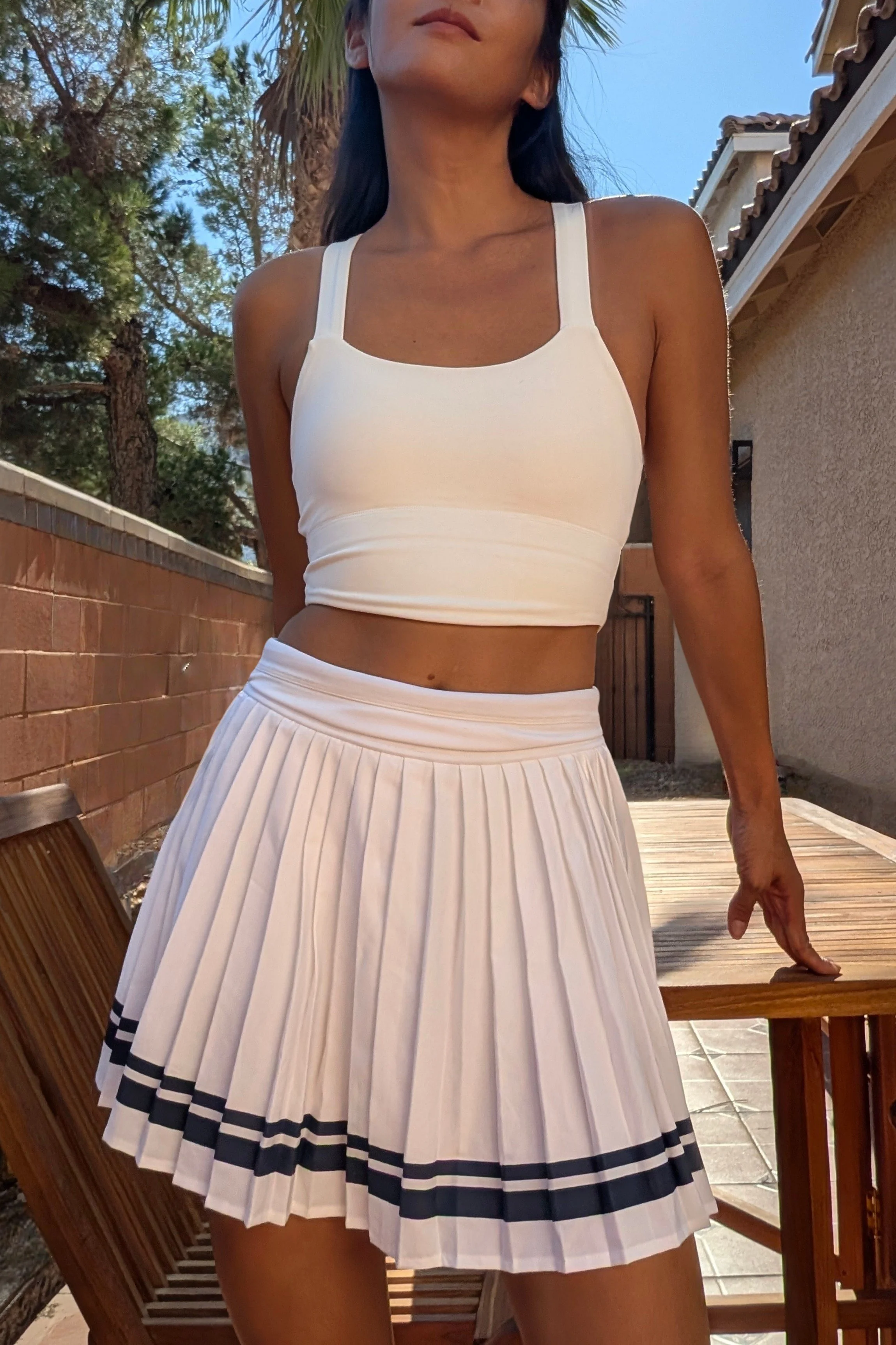 A woman outdoors wearing a white crop top and a white pleated skirt with black stripes at the hem, standing near a wooden chair, with trees and house exterior in the background. GFE Las Vegas, dinner date, escort, Scarlett Song Escort,