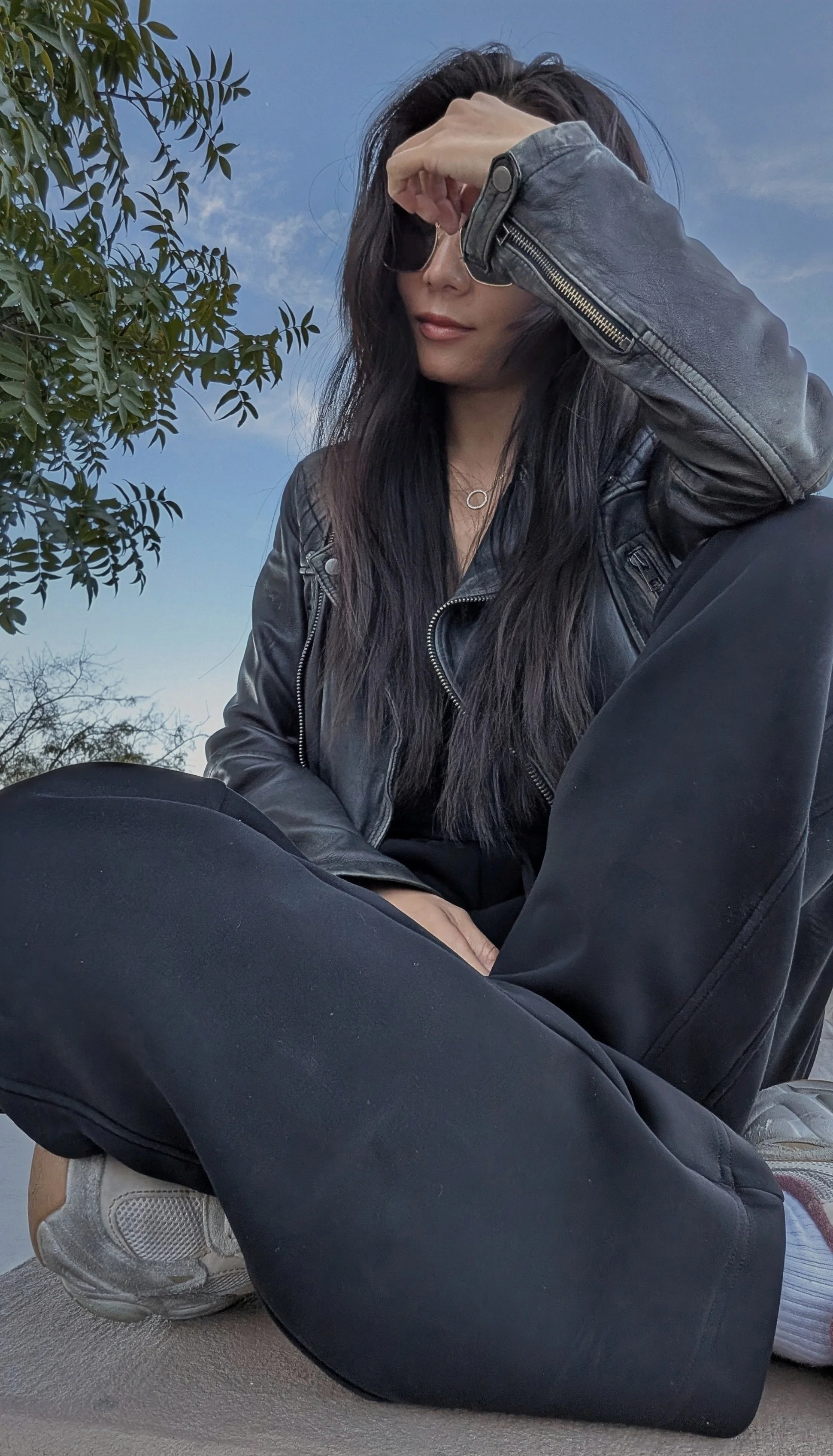 A woman with long dark hair sitting outdoors in a black leather jacket and black pants. She is wearing sunglasses and holding her forehead with one hand, with trees and a clear sky in the background. GFE Las Vegas, dinner date, escort, Scarlett Song 