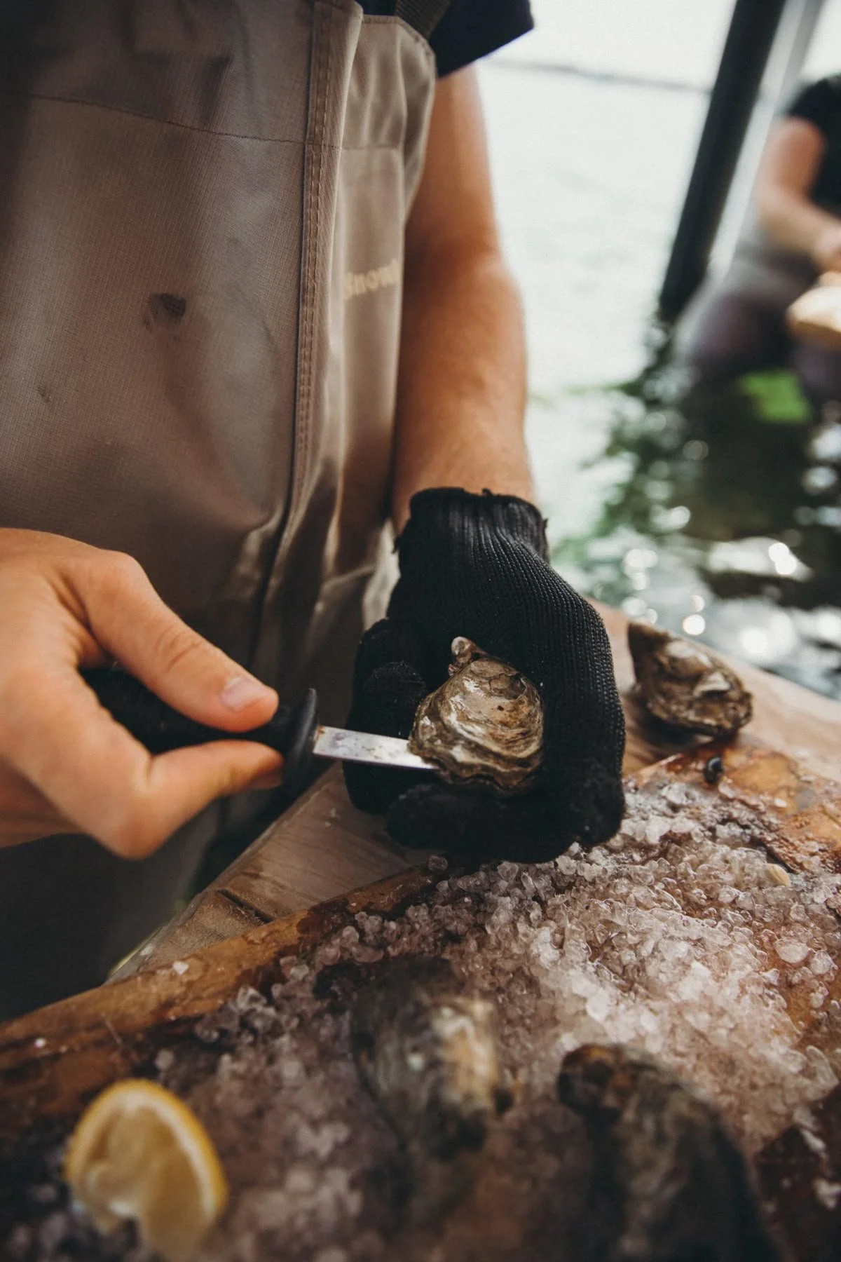 A person wearing black gloves shucking an oyster over a bed of ice on a wooden surface. There are lemon wedges on the ice, and water and another person in the background.
