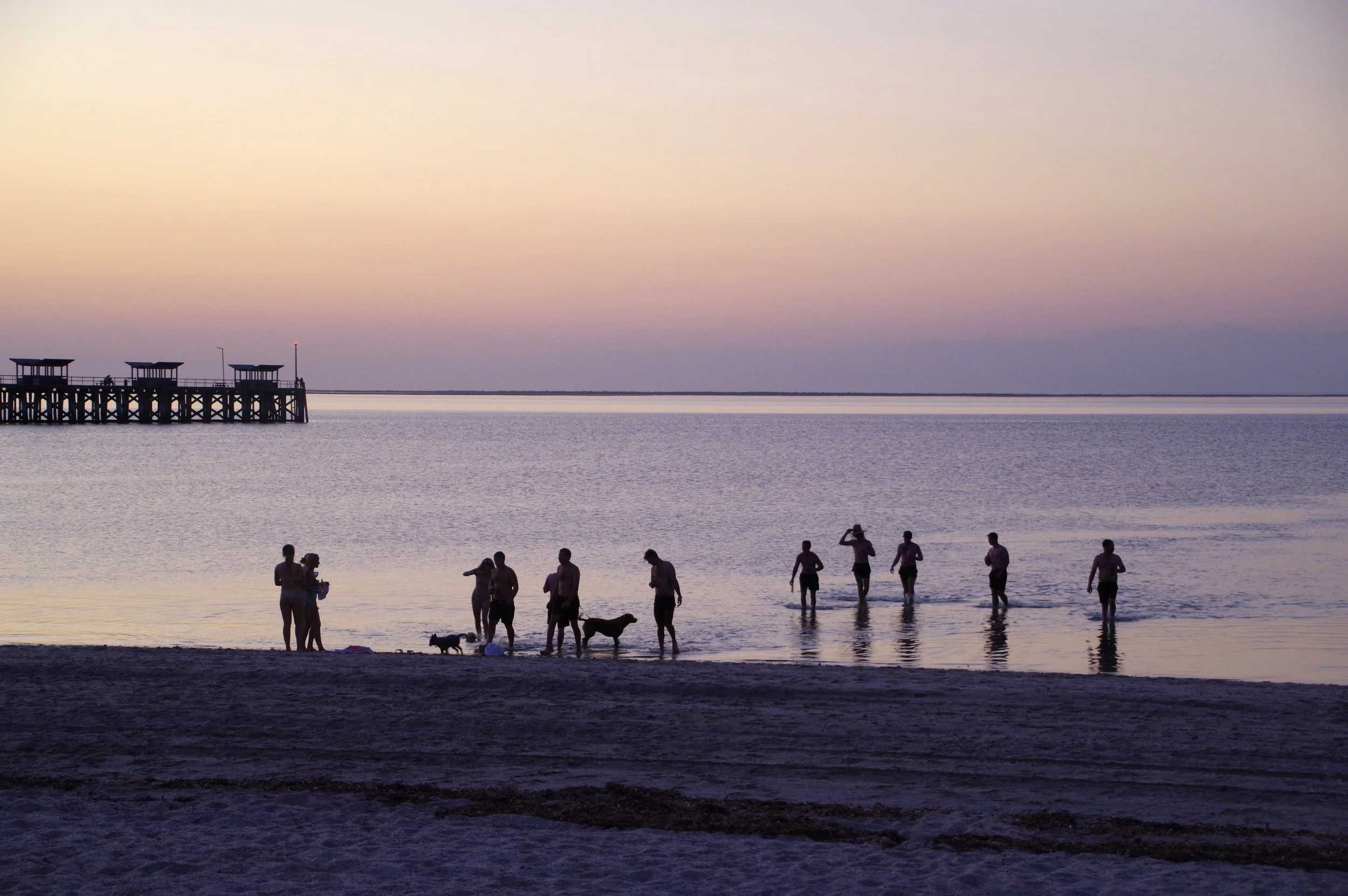 People wading in the ocean at sunset, with a pier in the distance.