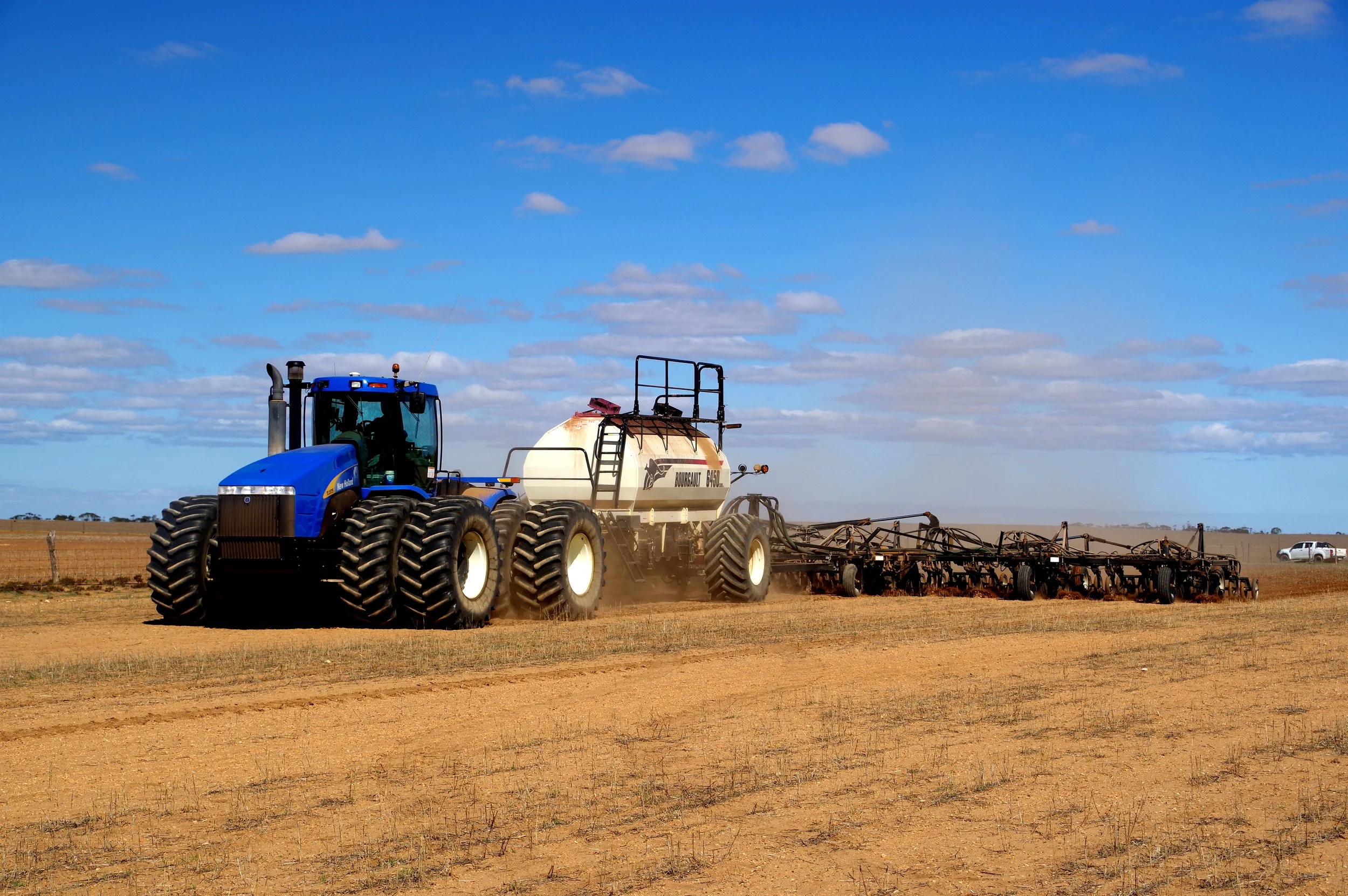 A large blue tractor pulling a white tank and other farming equipment across an open, flat, farm field under a partly cloudy sky.