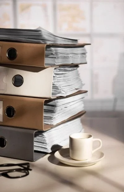 Stack of five binders filled with papers on a desk, with a coffee cup on a saucer in front and a pair of glasses nearby.