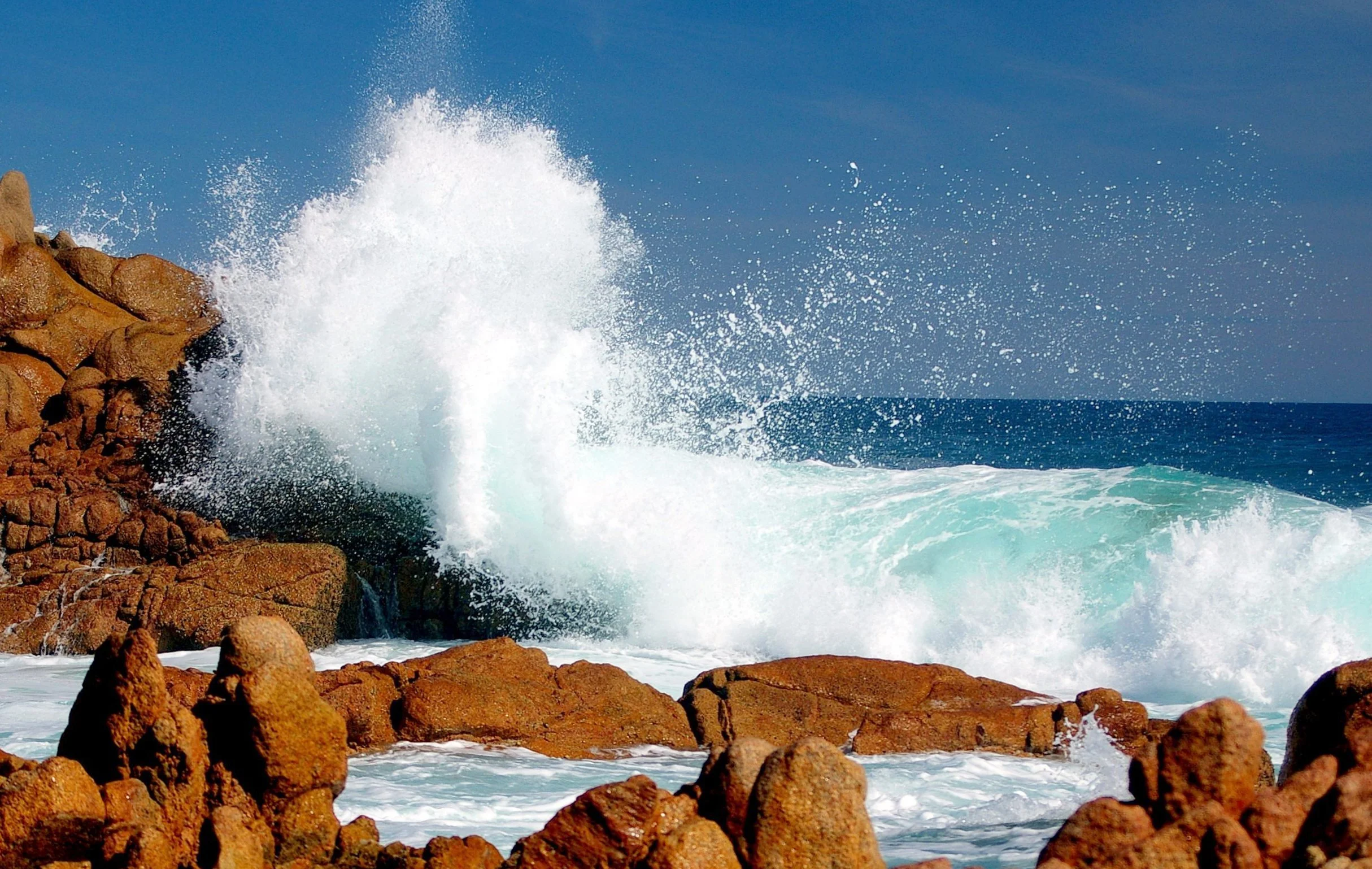 Large ocean wave crashing against brown rocky shoreline with a blue sky in the background.