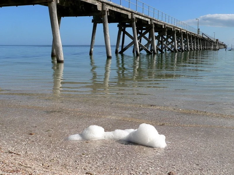 A wooden pier extending over calm, clear water with gentle waves reaching the sandy beach in the foreground; a small clump of white sea foam rests on the sand beneath a partly cloudy sky.