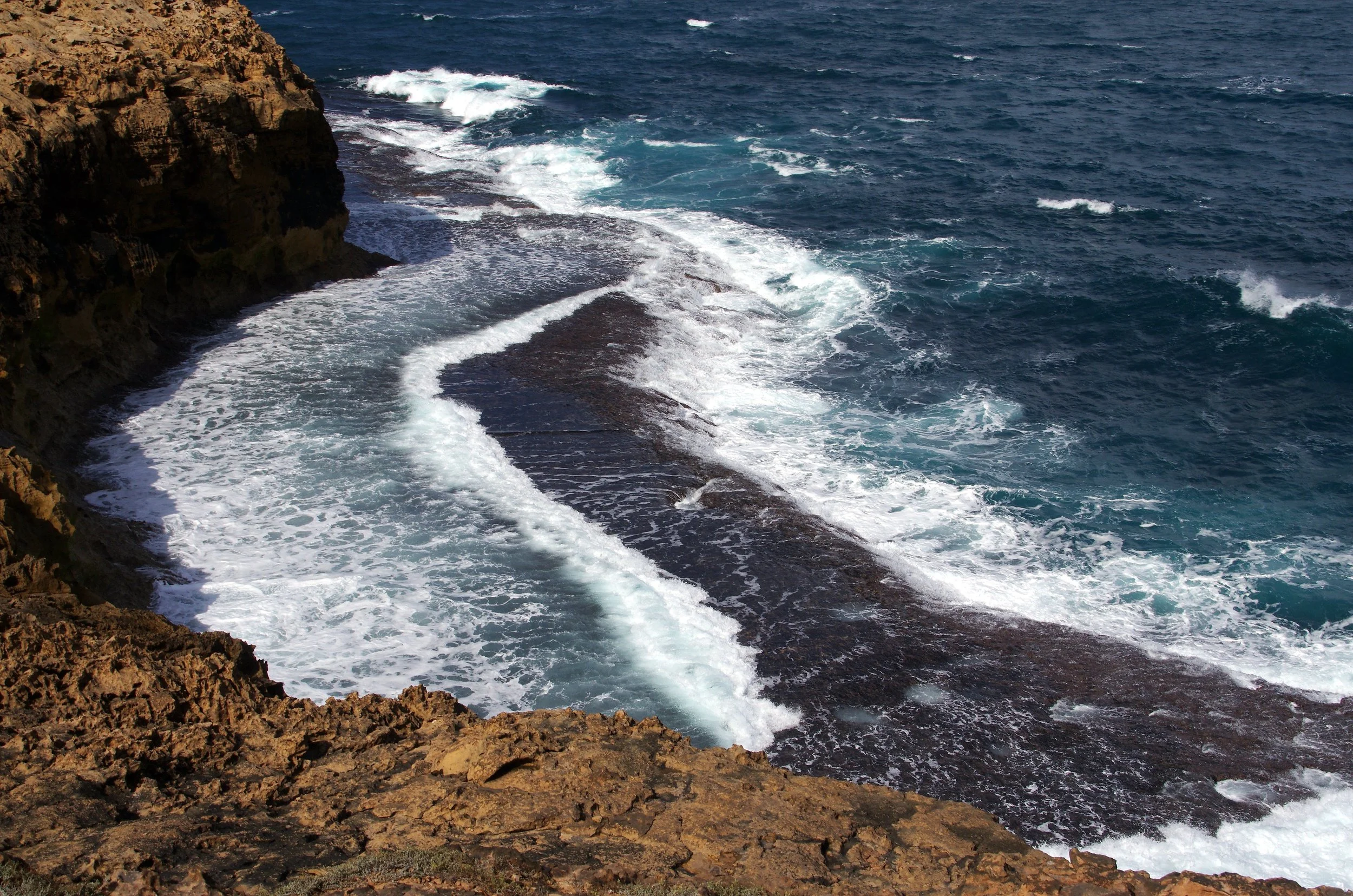 Cliffside view of the ocean with waves crashing against rocky shore, showing white foam and deep blue water.