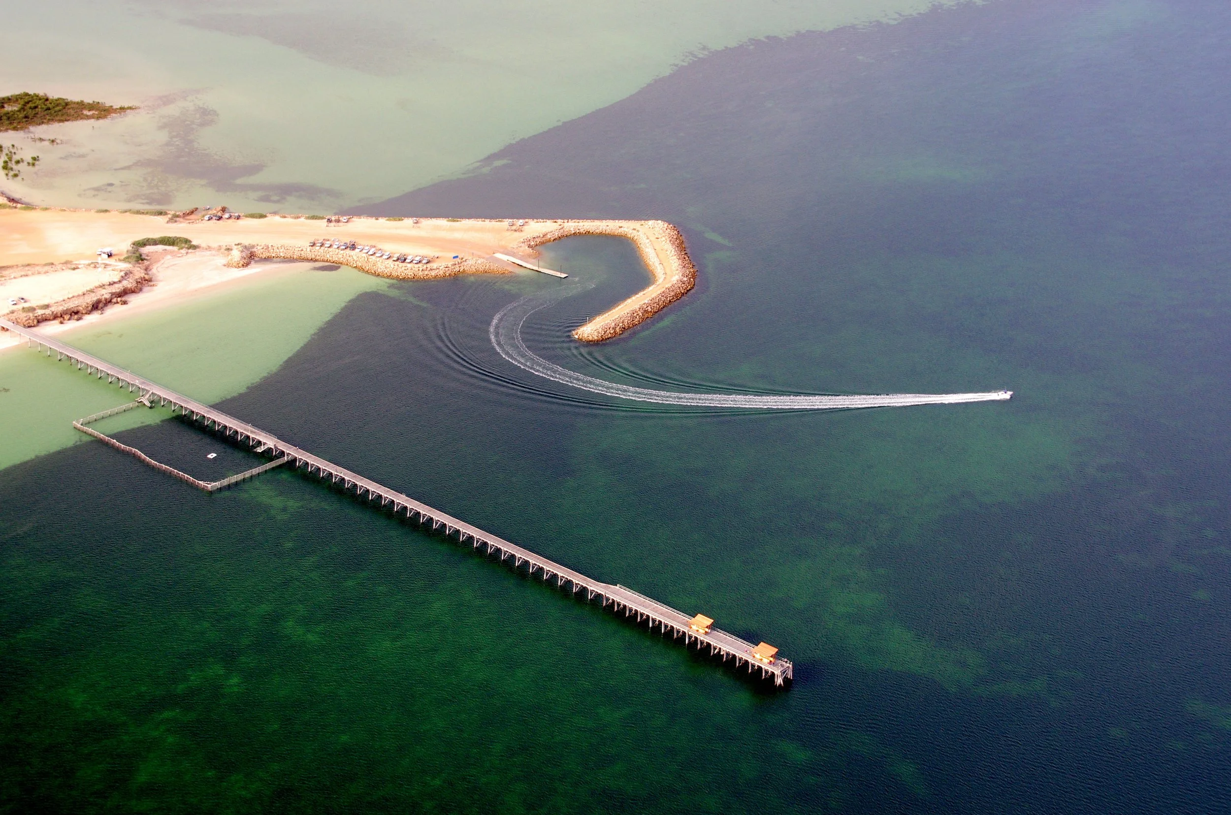 Aerial view of a pier extending into the water with boats creating waves near the breakwater, and a sandy area with parked cars and a small building.