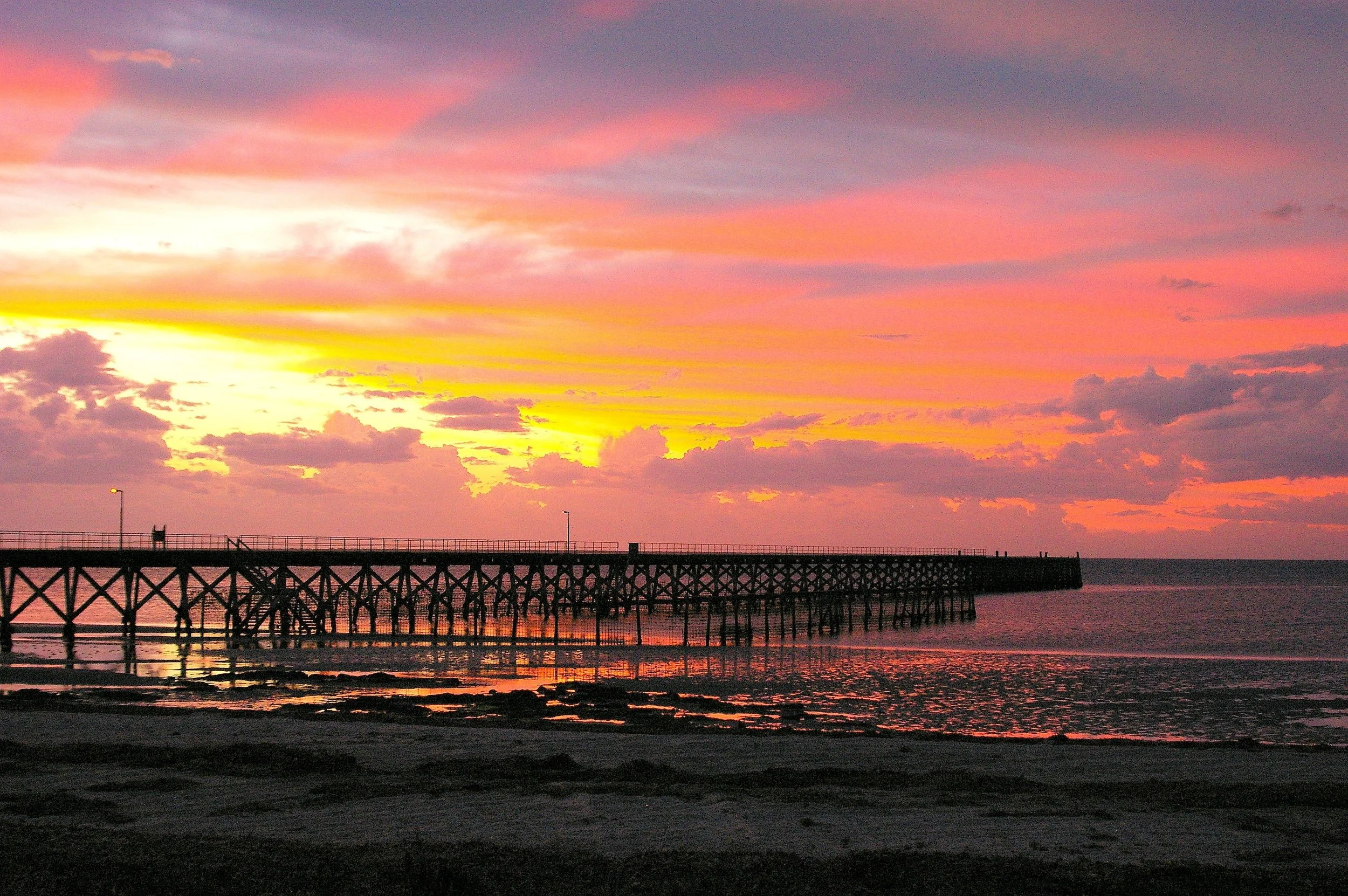 Sunset over a pier extending into the ocean with colorful pink, orange, and purple sky and calm water reflections.