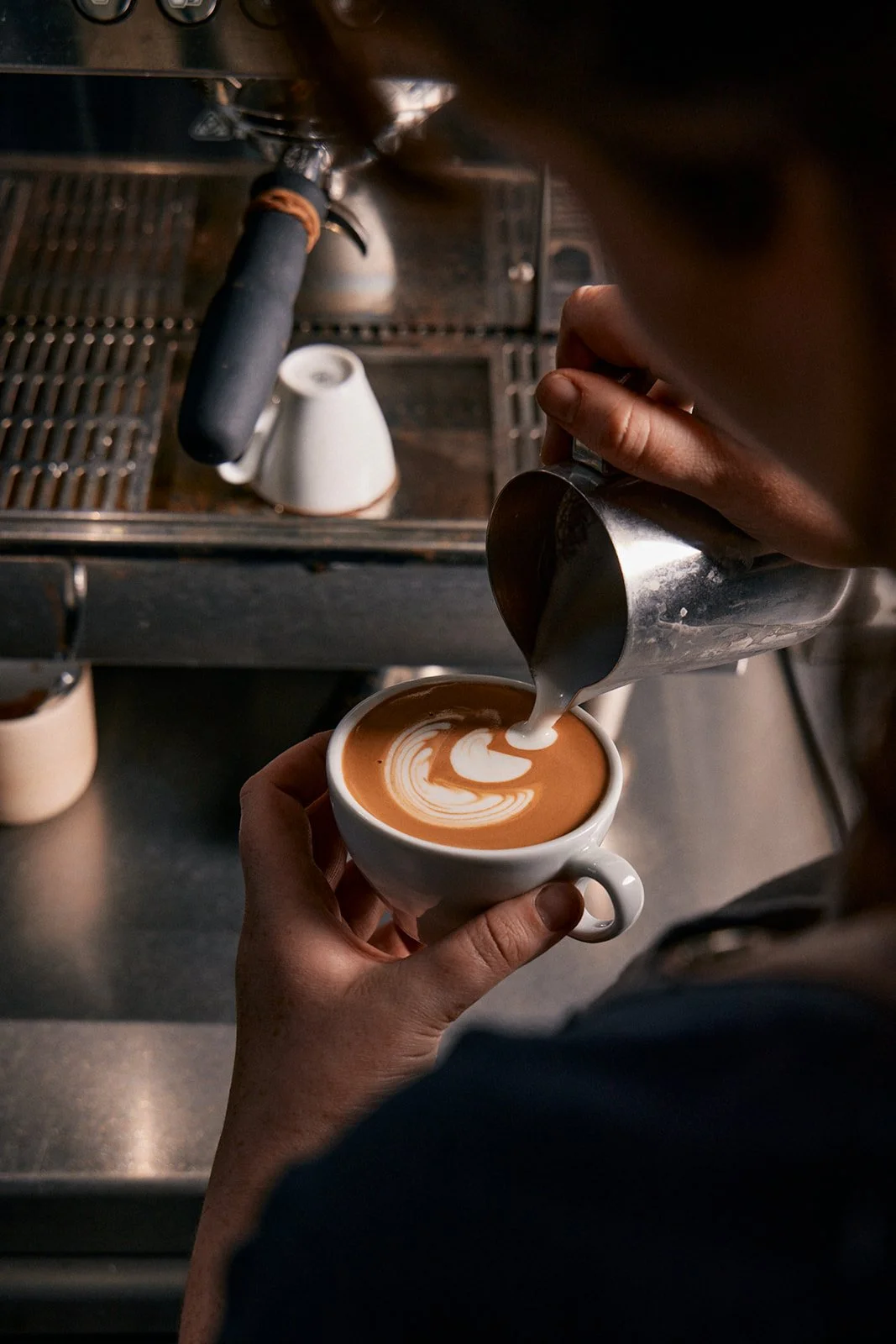 Empty dining room with a circular wooden dining table and six wooden chairs, decorated with small glass vases, under a modern elliptical ceiling light fixture, with a wooden floor and plain light-colored walls.