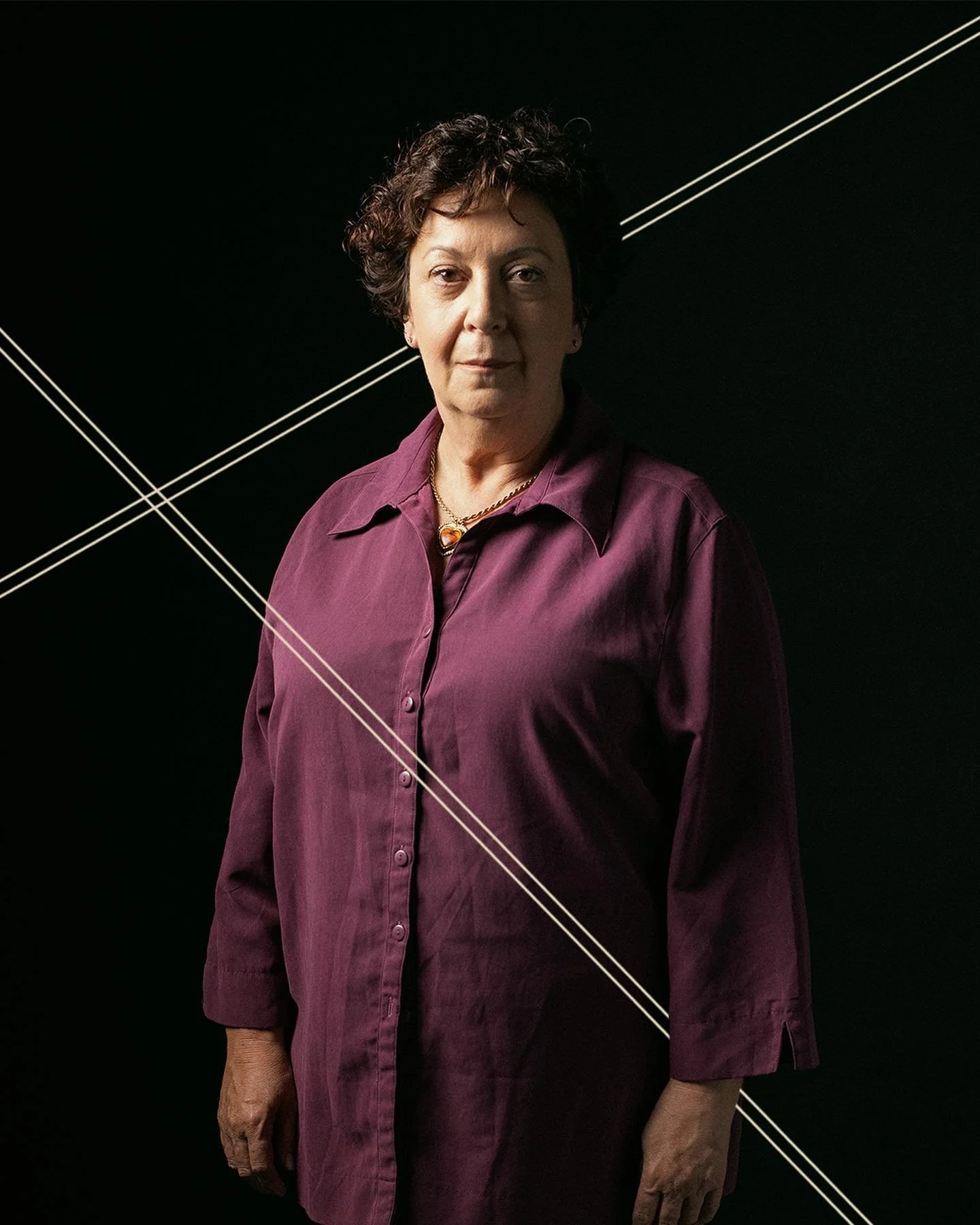 A woman with short, dark curly hair wearing a purple button-up shirt stands against a black background with thin white diagonal lines crossing behind her.