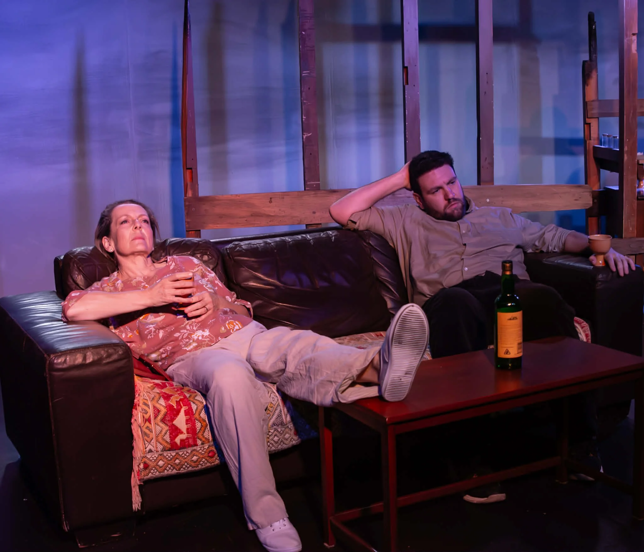 Two people sitting on a dark brown leather couch, relaxing. The woman, wearing light-colored pants and a floral shirt, is holding a glass and has her legs resting on the table. The man, in a beige shirt, appears to be lost in thought, holding a cup. 