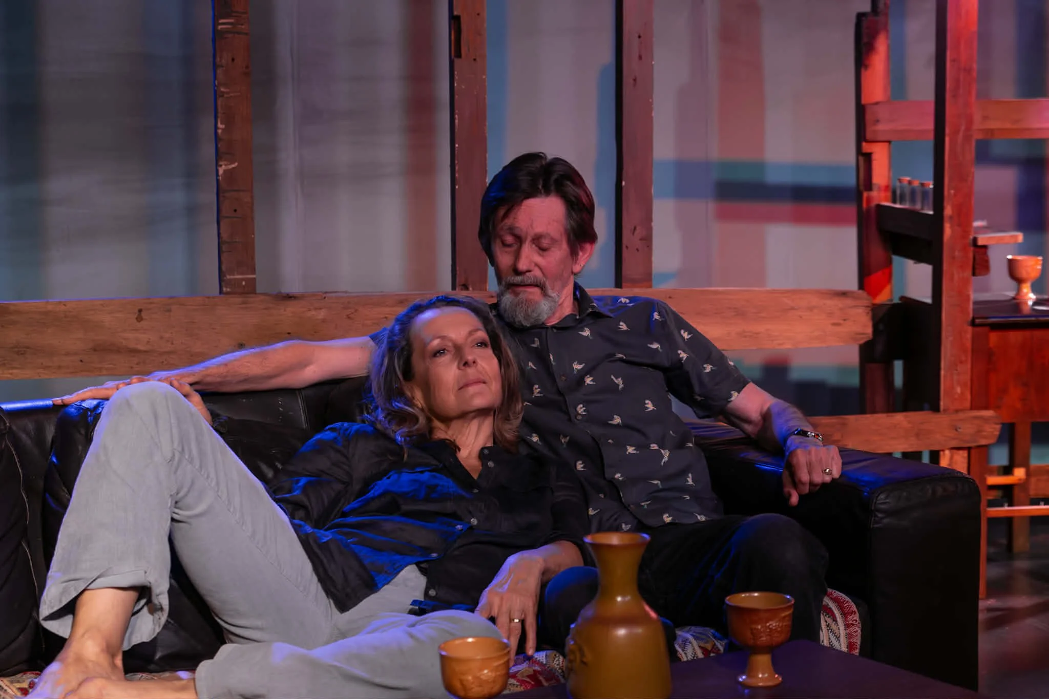 Two people sitting on a black leather sofa in a cozy room with wooden furniture, including a coffee table with two pottery cups and a vase, and a wooden shelf with small items. The woman is reclining with one arm stretched out along the back of the s