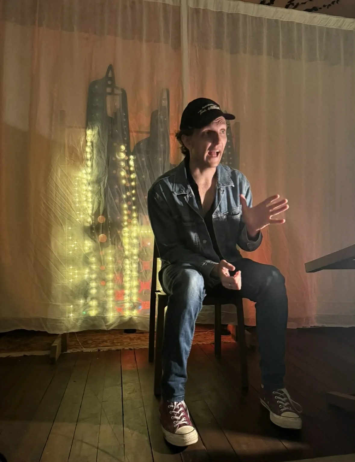 A young man sitting on a chair, wearing a denim jacket, black shirt, jeans, and red sneakers. He appears to be speaking or gesturing with his right hand. Behind him is a soft orange curtain with vertical sheer panels, and a lit-up decorative backdrop