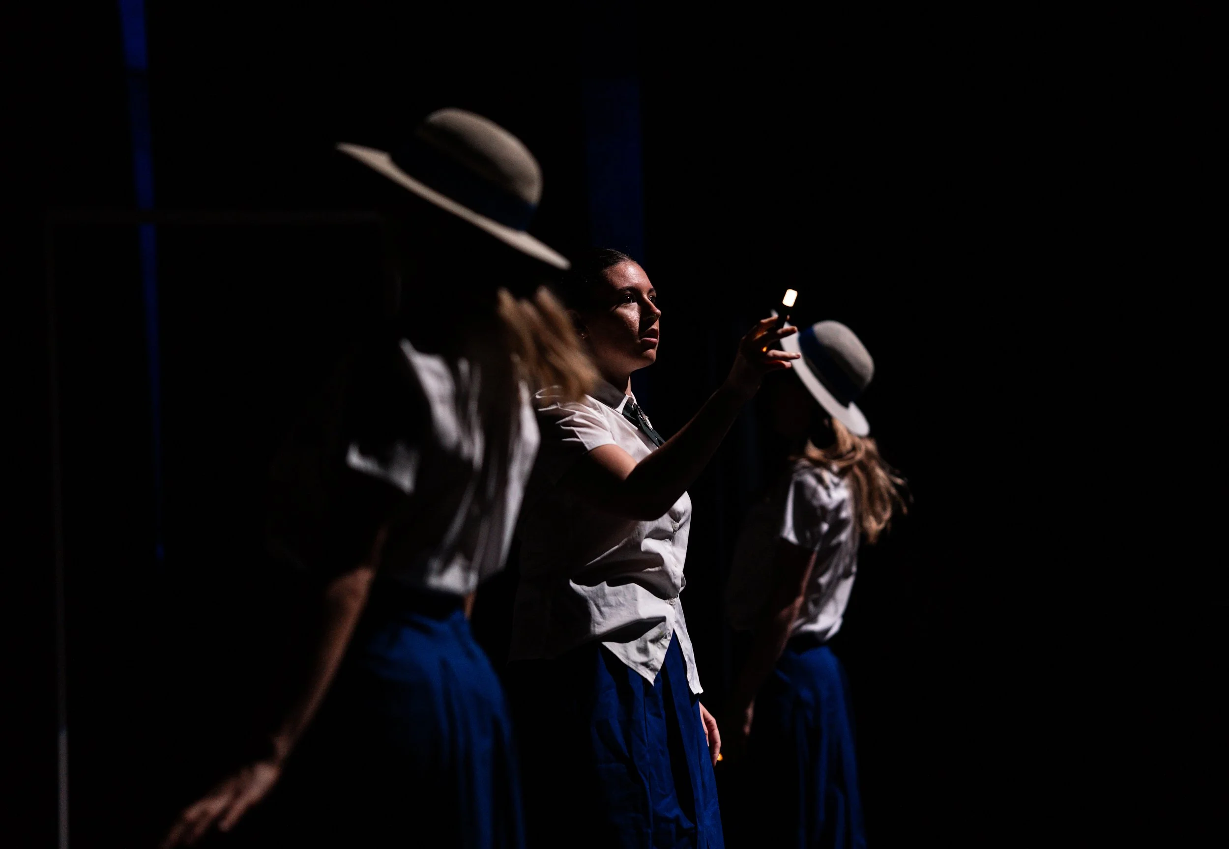 Three women in white shirts and blue skirts wearing wide-brimmed hats stand in a dark space, with one woman illuminated, holding a glow-in-the-dark marker.