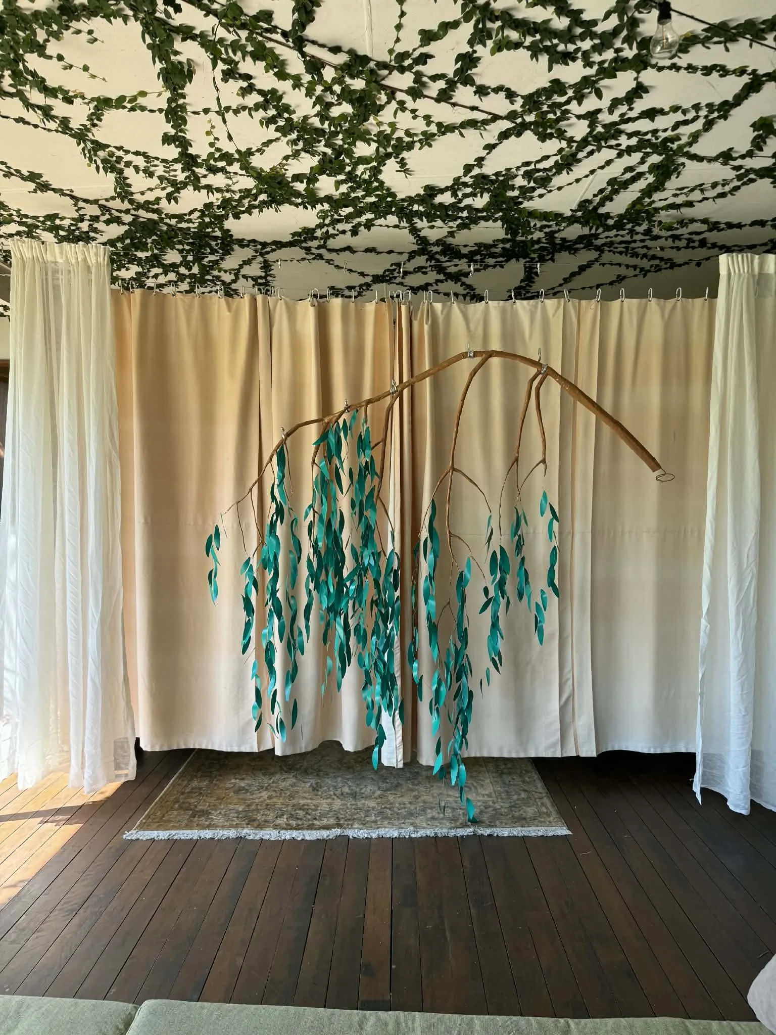 Indoor setting with beige curtains, decorative hanging branch with teal leaves, and greenery on the ceiling.