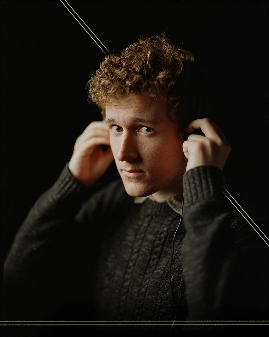 A young man with curly hair wearing a dark sweater, looking at the camera while adjusting earphones against a black background.