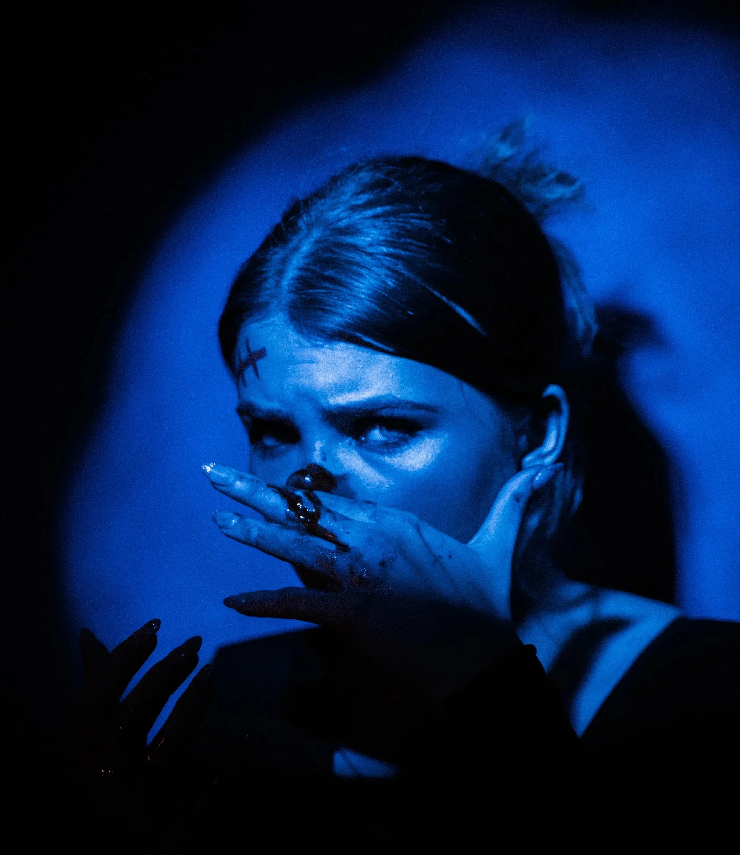 Woman with dark hair and black makeup, covering her mouth with her hand, looking intensely at the camera against a dark background with blue lighting.
