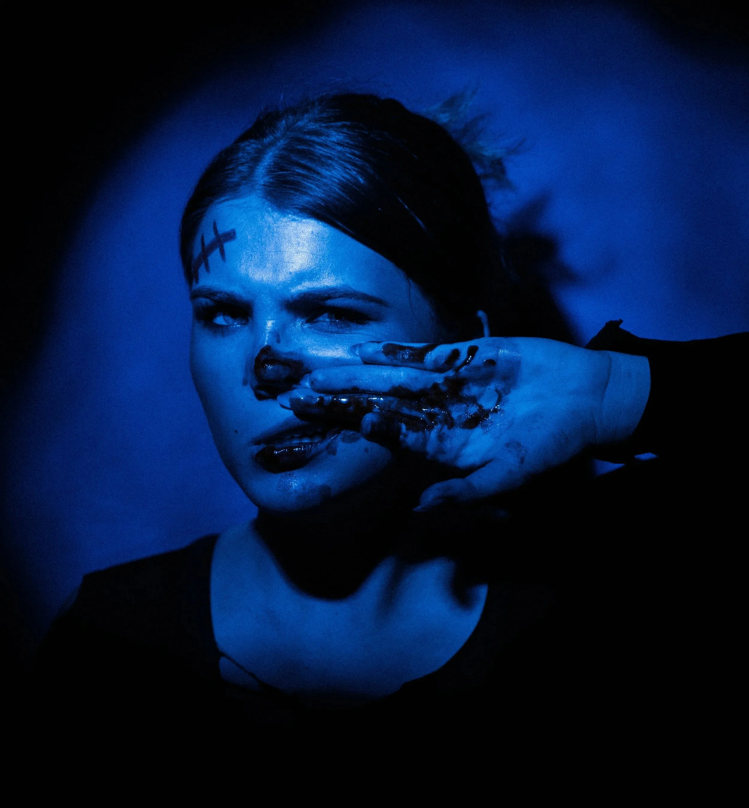 A woman with dark hair and makeup holding her hand to her face, covered in dark paint or mud, illuminated by blue lighting with a dark background.