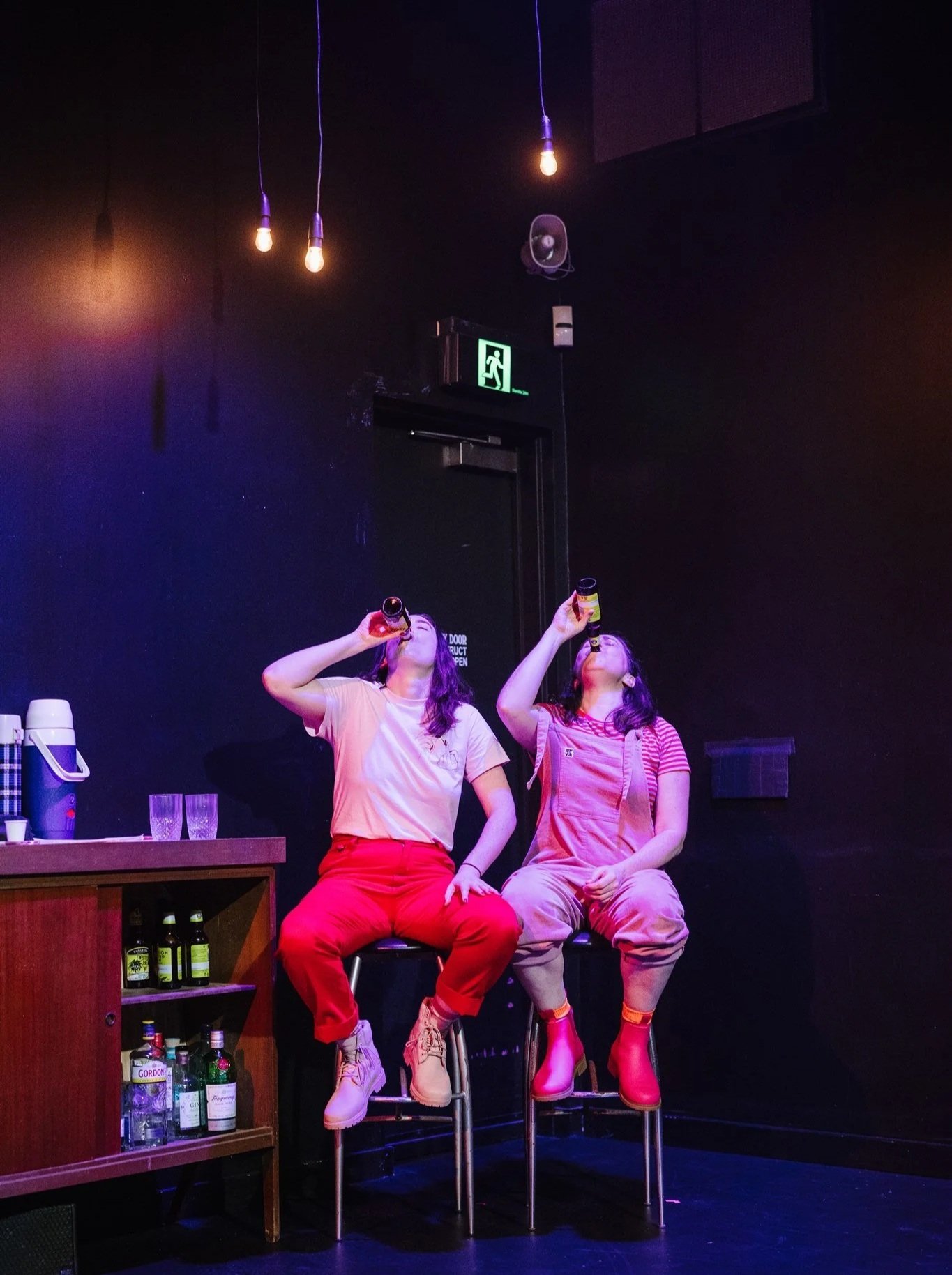 Two women sitting on bar stools drinking bottles of beer in a dimly lit room with hanging light bulbs and a black wall.