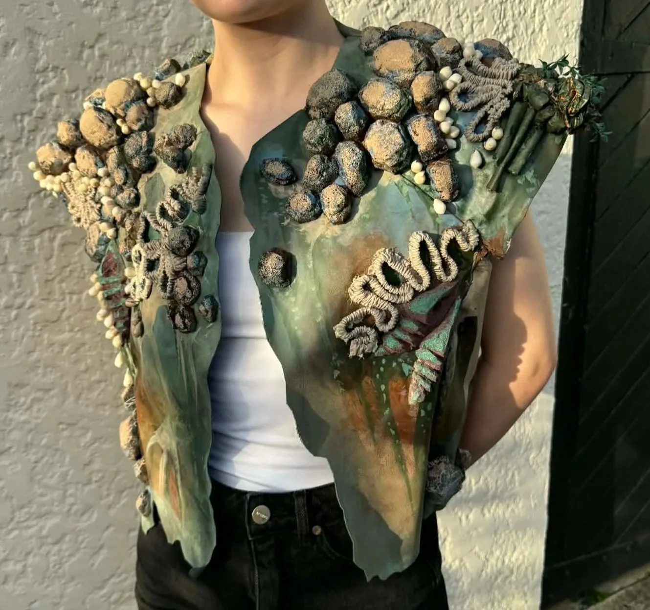 Person wearing a jacket decorated with natural elements like shells, rocks, and plants, standing outdoors against a textured wall and black wooden structure.