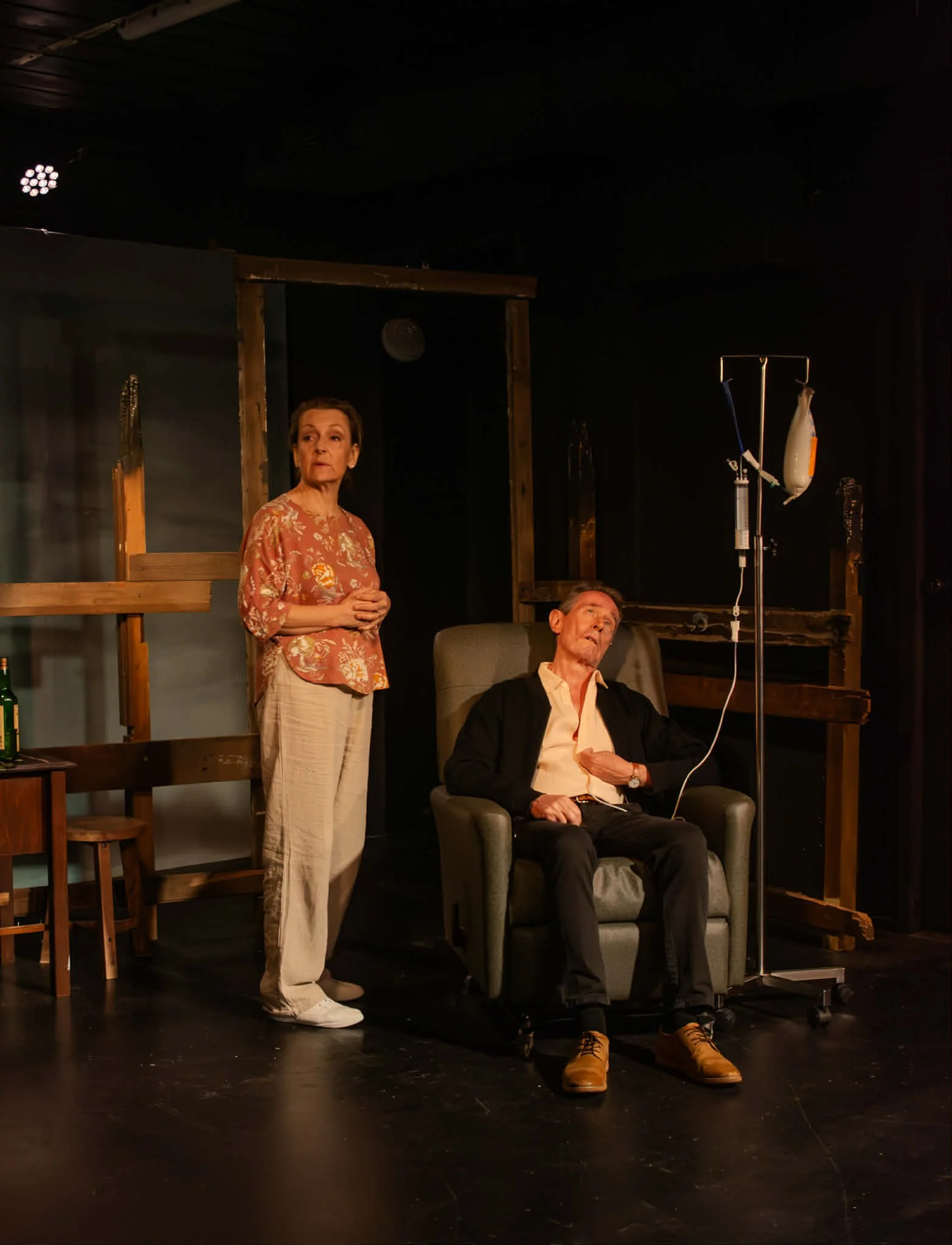 A theater scene with two actors, one woman standing and a man sitting in a chair with an IV drip, set against a dark background with wooden props.