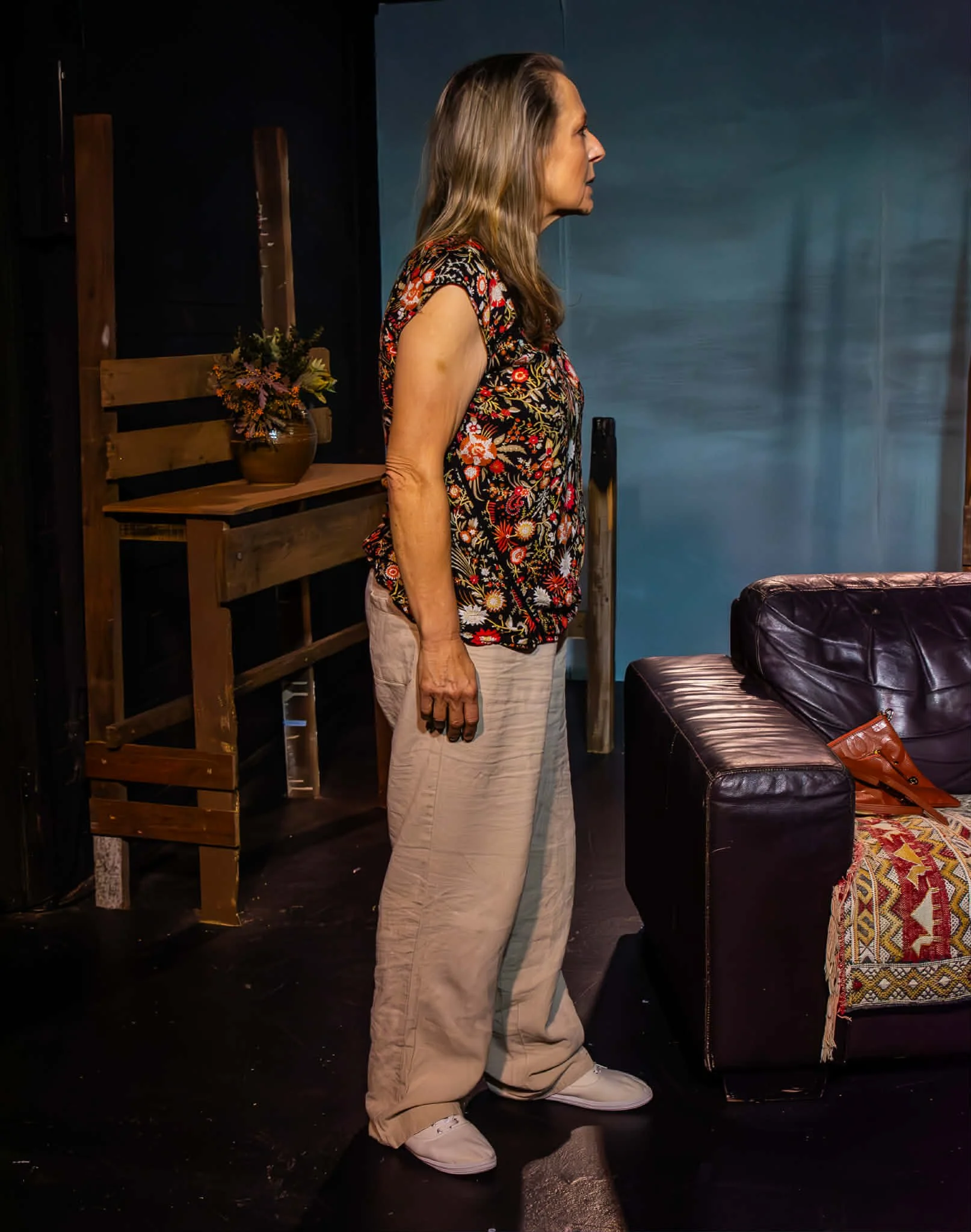 A woman standing indoors with her face in profile, wearing a floral top and beige pants, near a dark leather couch.