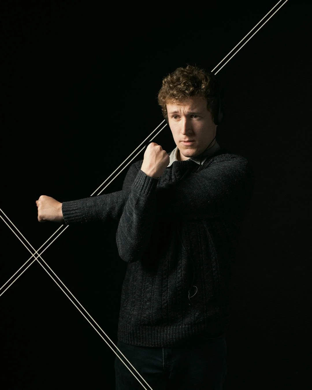 A young man with curly hair wearing a black sweater and headphones, standing against a black background with white geometric lines, making a fist with his right arm.
