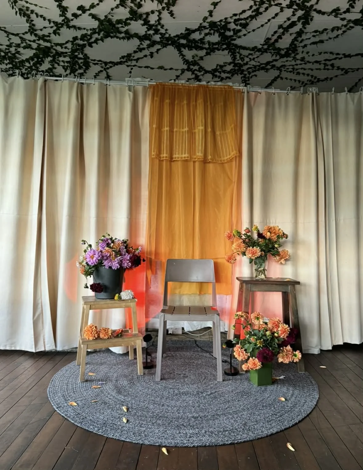 A small stage setup with beige curtains, an orange sheer backdrop, two wooden tables with flower arrangements, a gray chair, a gray rug, and leaves scattered on the wooden floor.