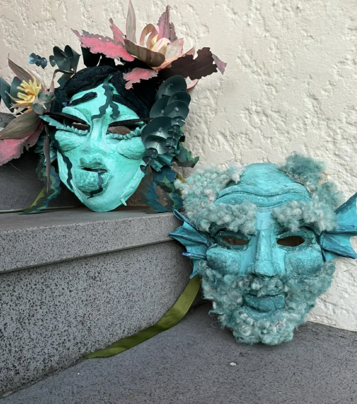 Two elaborate masks resting on a concrete step; one mask features a face with black and teal paint, pink and cream flowers and leaves around the head, and striped black and teal details; the other mask is mostly teal with a fluffy texture, small horn