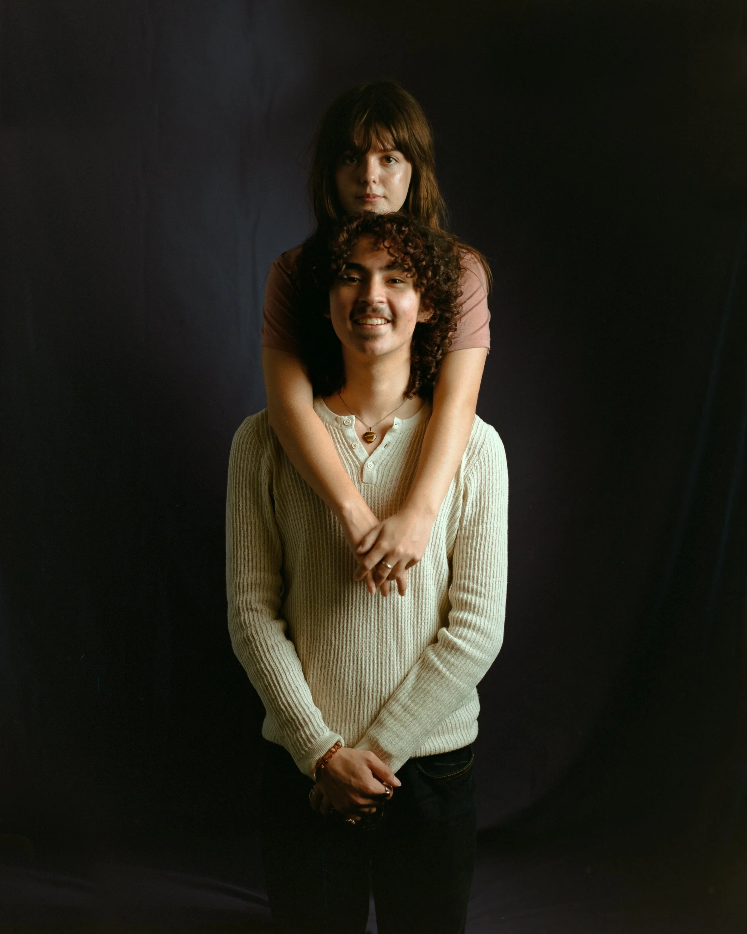 A young woman with brown hair and a young man with curly hair posing together, with the woman standing behind and embracing the man, both smiling at the camera against a dark background.