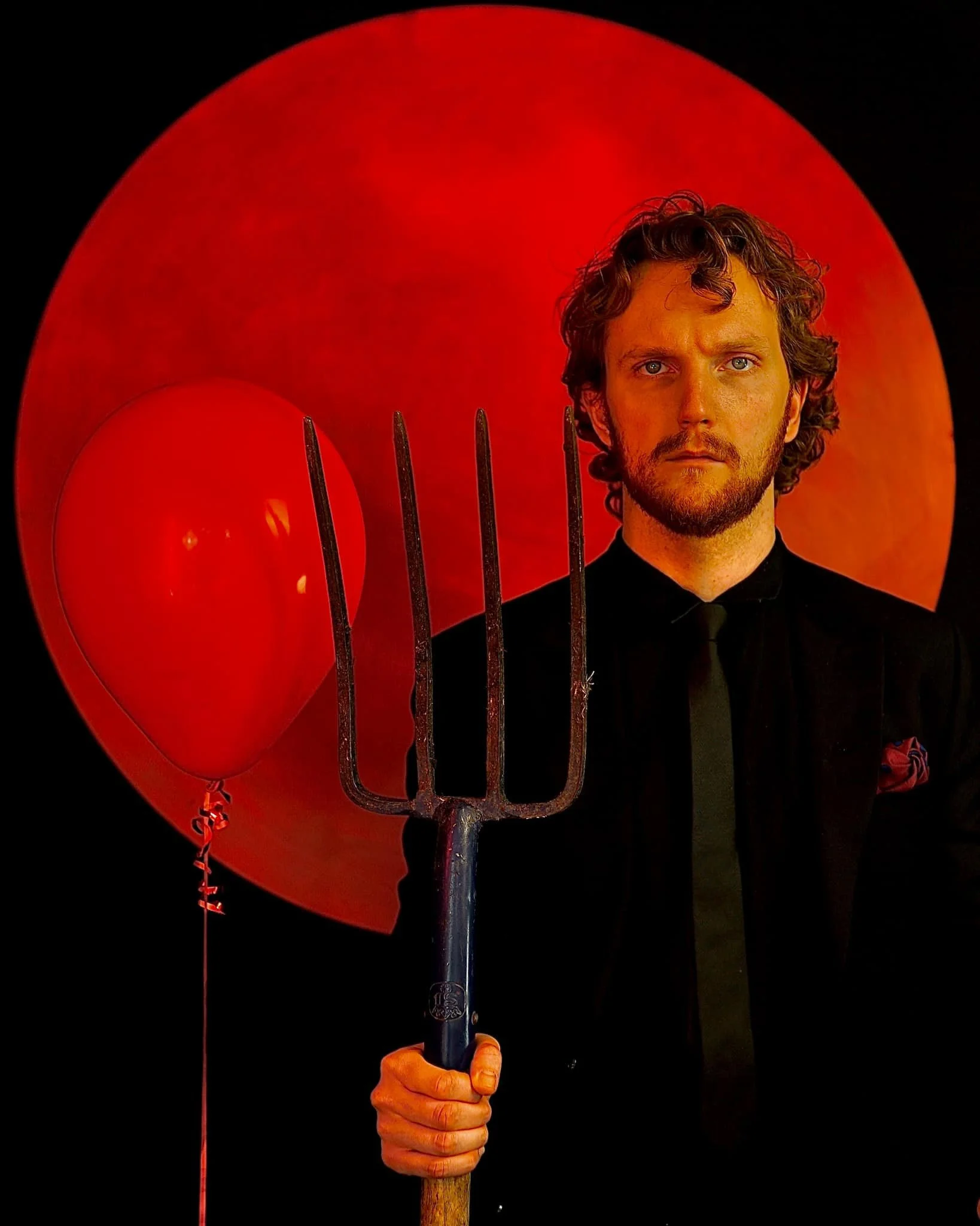 A man with curly hair and a beard, dressed in a black suit, holding a pitchfork, standing in front of a large red circle with a red balloon beside him.