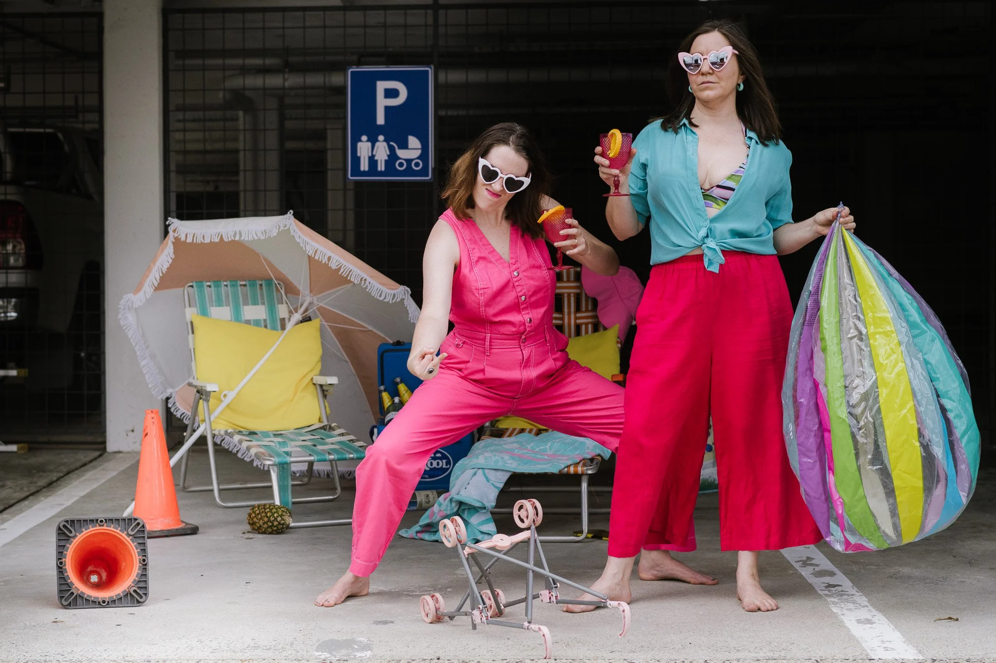 Two women wearing bright pink pants and colorful tops standing by a parking lot under a parking sign, with beach accessories and drinks in hand, next to a beach chair, umbrella, and toy stroller.