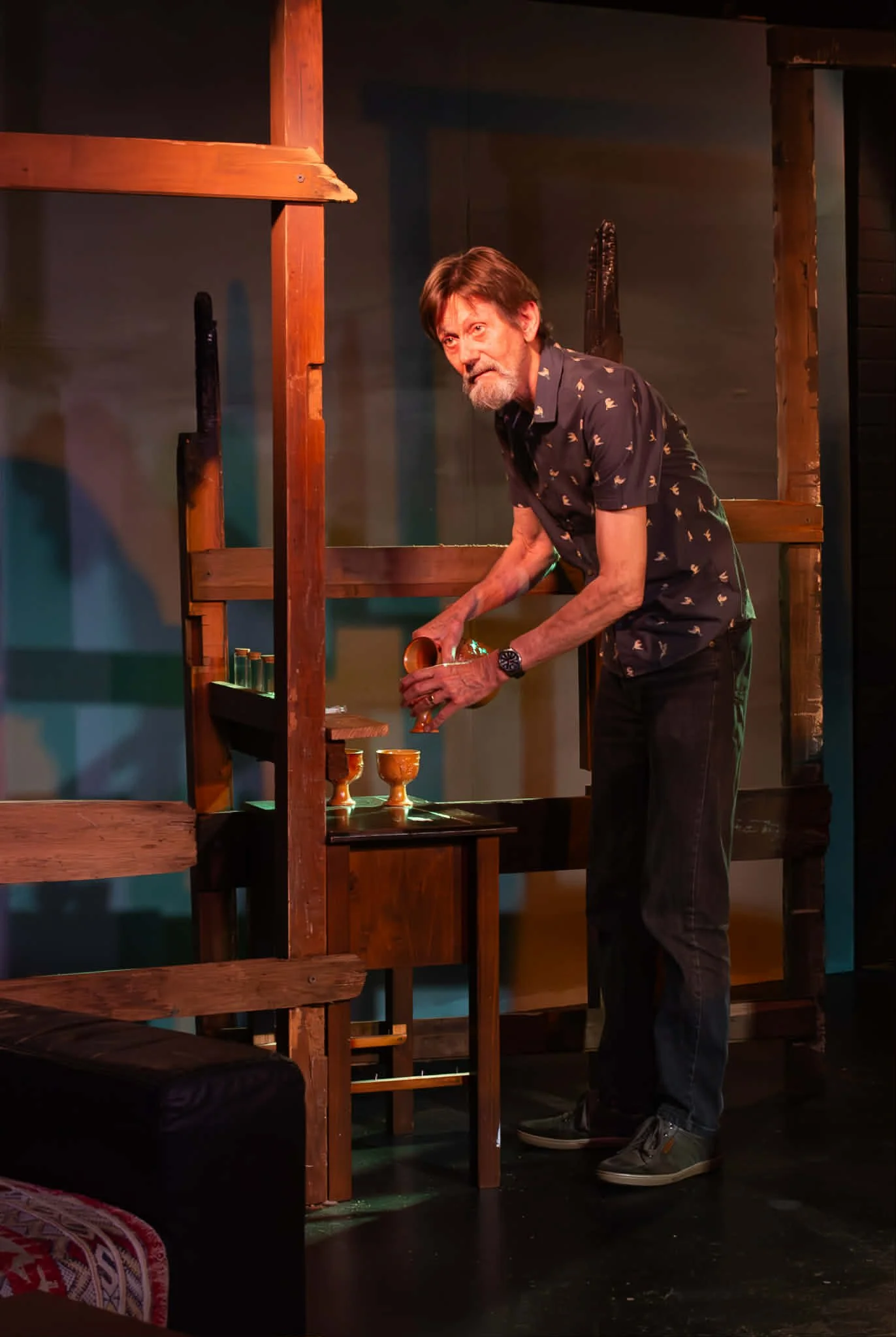 A man with gray hair and beard, wearing a dark patterned shirt and black pants, sets a goblet on a wooden shelf during a stage performance.