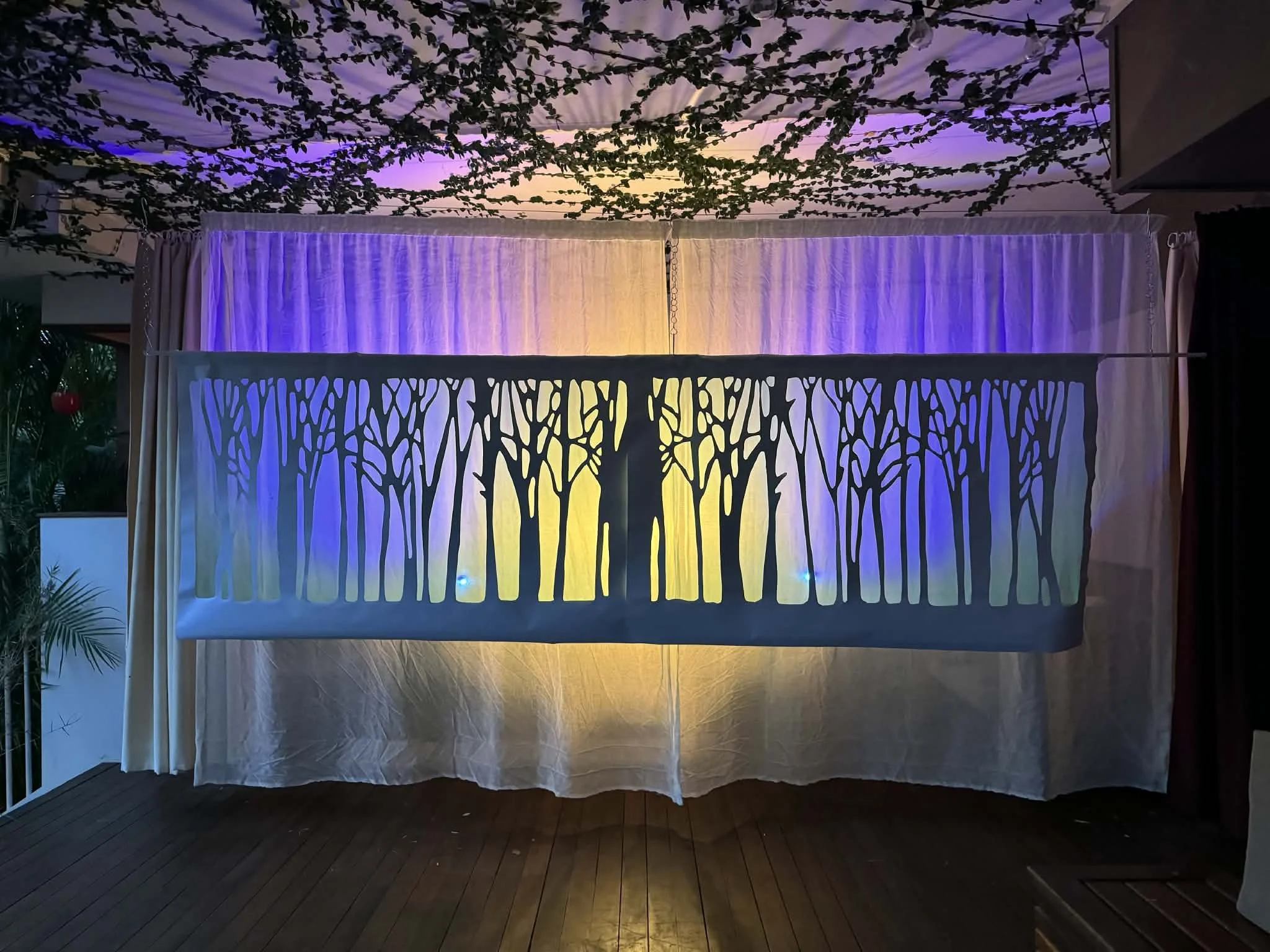 Fabric backdrop with a cut-out tree silhouette, illuminated by colorful lights, set against a ceiling with hanging green foliage and decorative string lights.