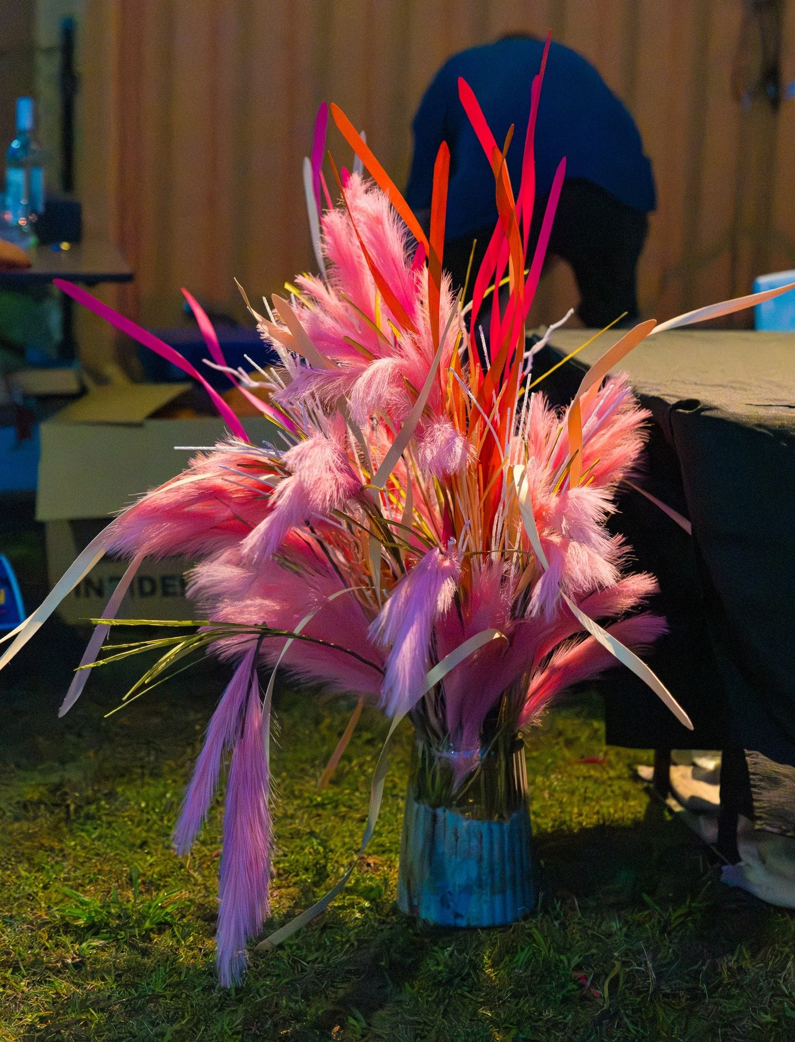 A colorful bouquet of pink, orange, purple, and white feathers and long, thin leaves in a small, cylindrical glass vase on grass.