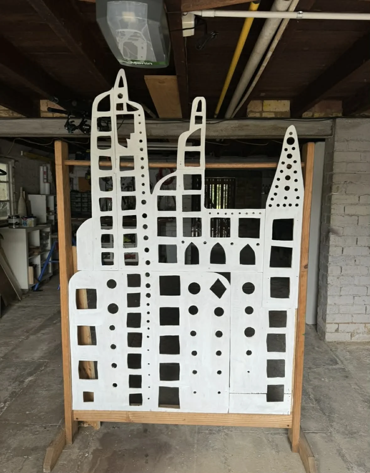 A large wooden cutout with an abstract city skyline design, painted white with various cutout shapes, stands on a wooden frame inside a workshop.