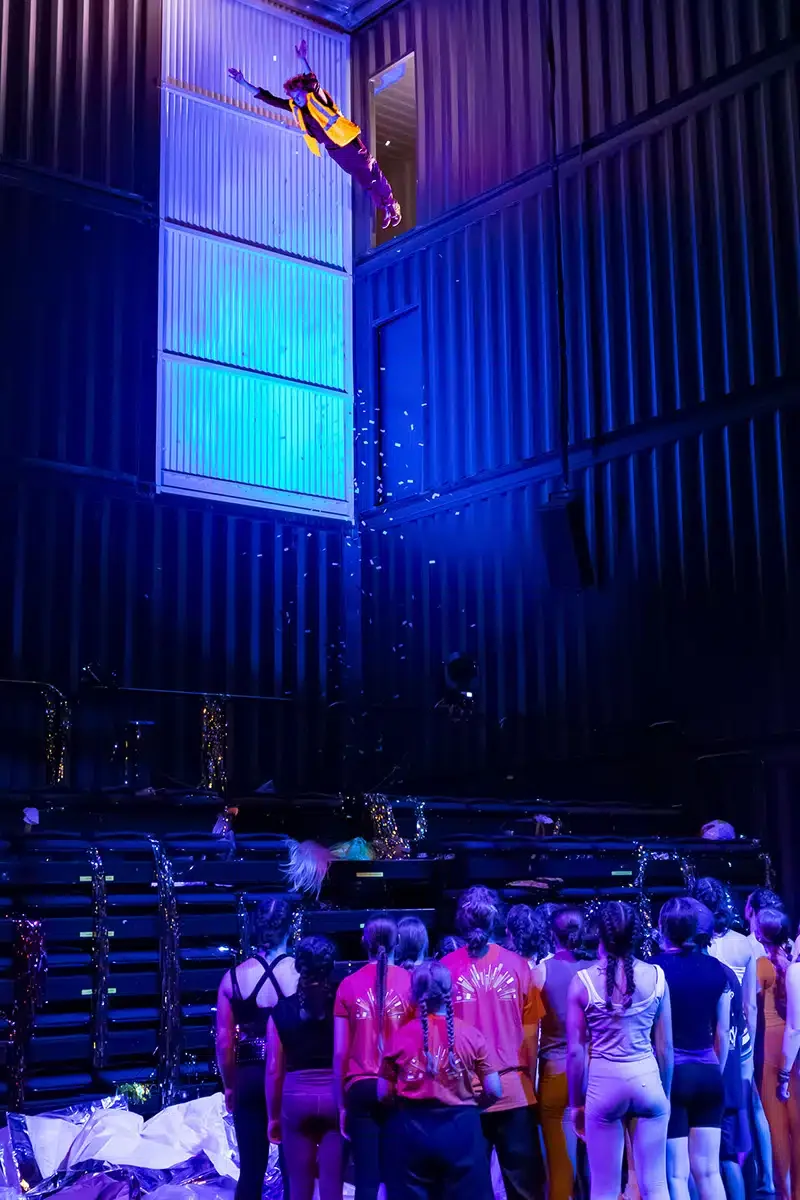 A person in yellow and purple attire being thrown or falling from a height into a pile of people on a stage with dark walls, under blue and purple lighting.
