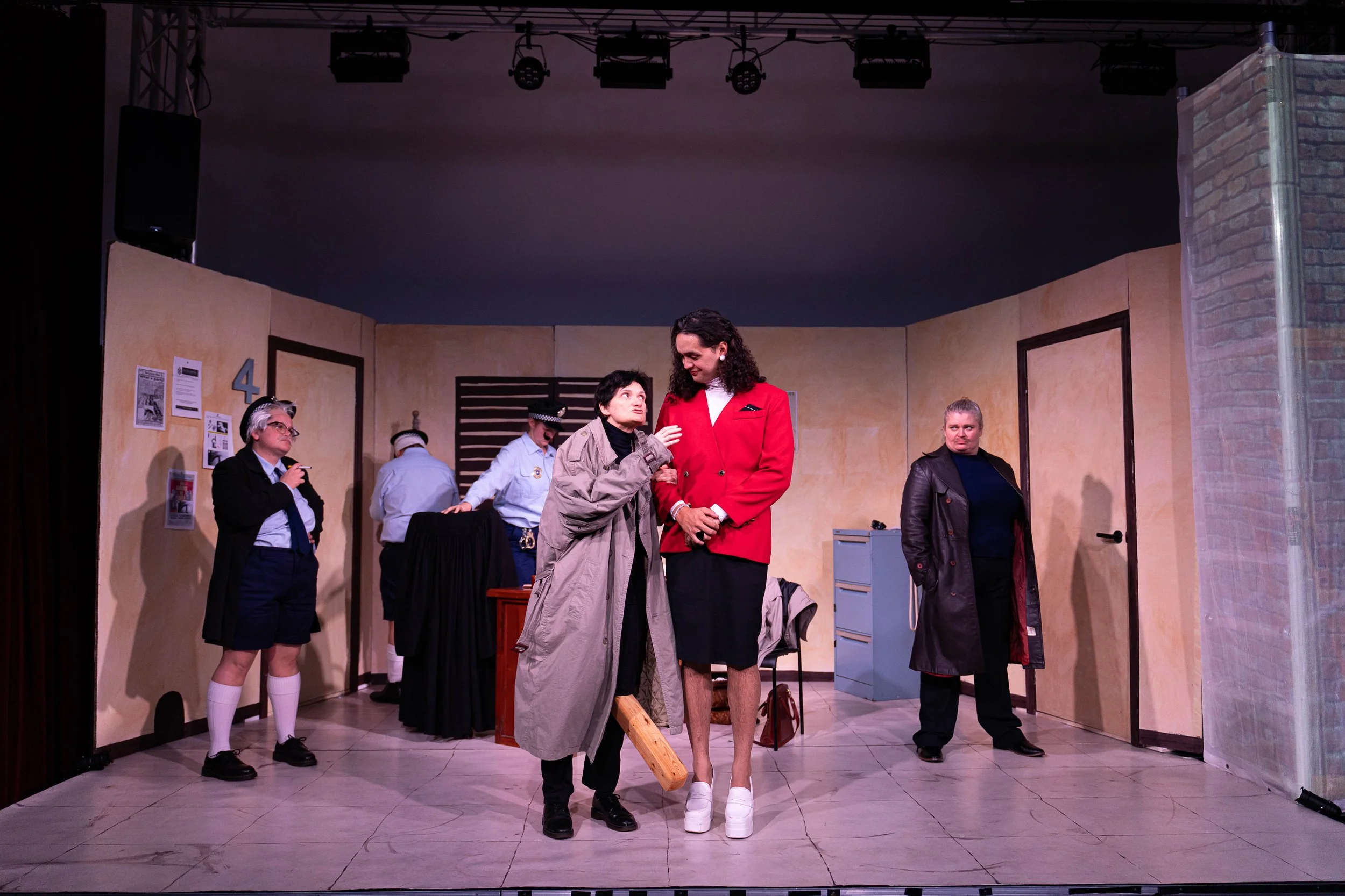 Actors on stage in a theatrical play set resembling an office, with five women, some in police uniforms, others in casual or professional attire, engaging in a scene.