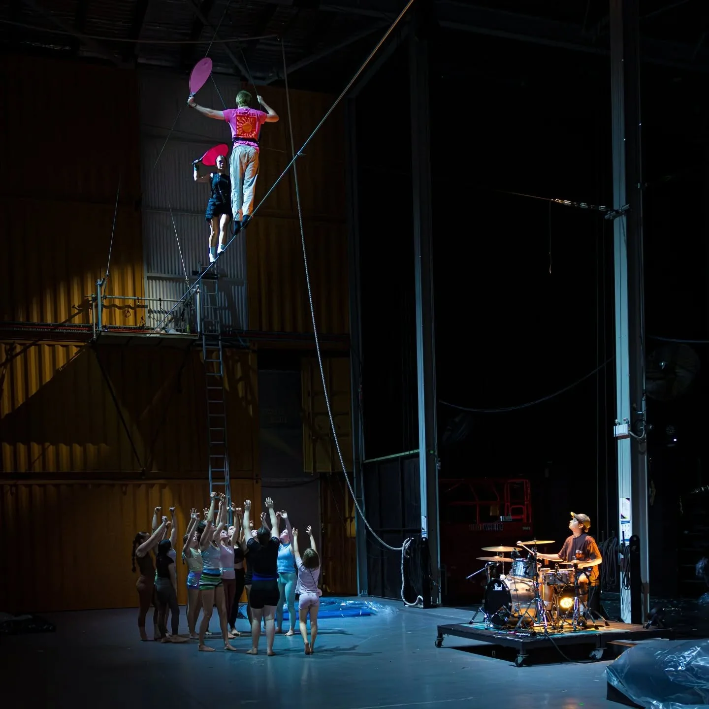 Circus performers practicing on a tightrope above a group of children with their arms raised, a drummer playing nearby, and a warehouse-like setting.