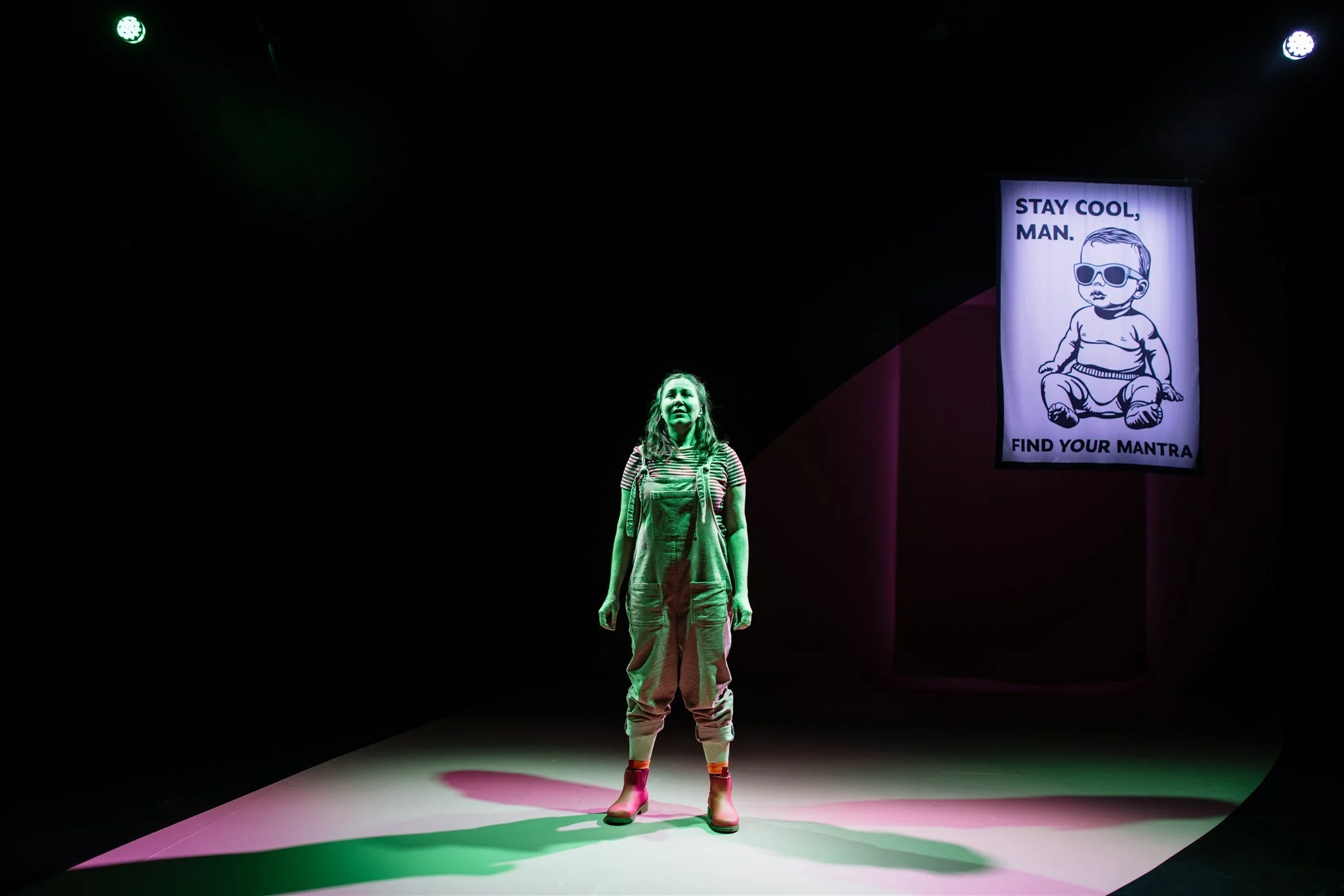 A woman standing on stage under green lighting with a dark background. There is a sign behind her that depicts a child in sunglasses with the text, 'Stay cool, man. Find your mantra'.