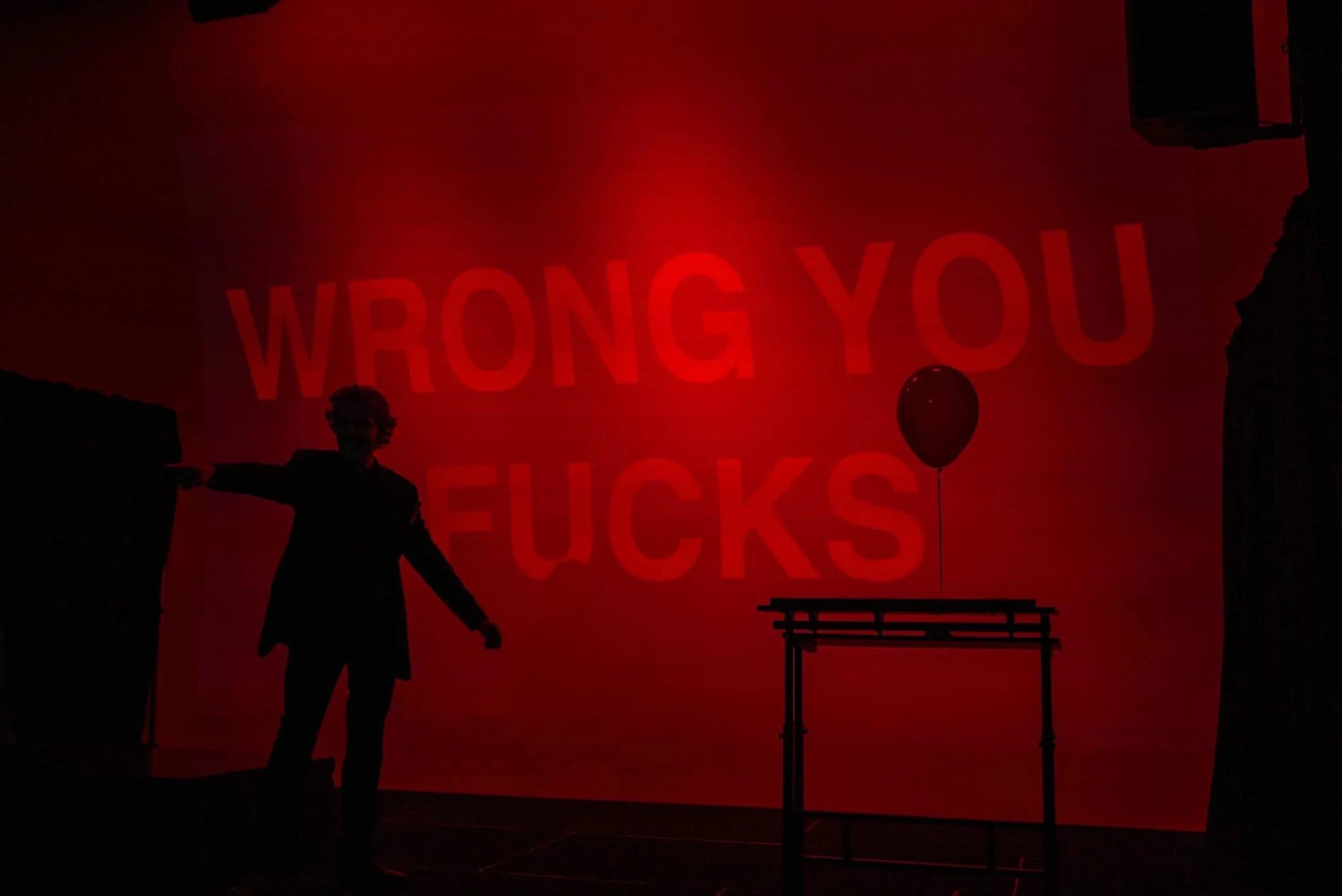 Silhouette of a person pointing with a large projected message reading 'WRONG YOU FUCKS' on the wall behind and a single balloon attached to a table.