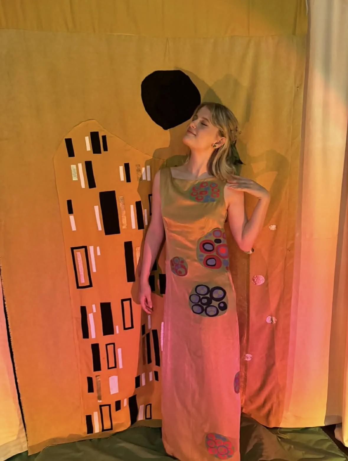 A woman in a colorful, patterned dress stands against an orange and black abstract background, with her eyes closed and a content expression, touching her neck with one hand.